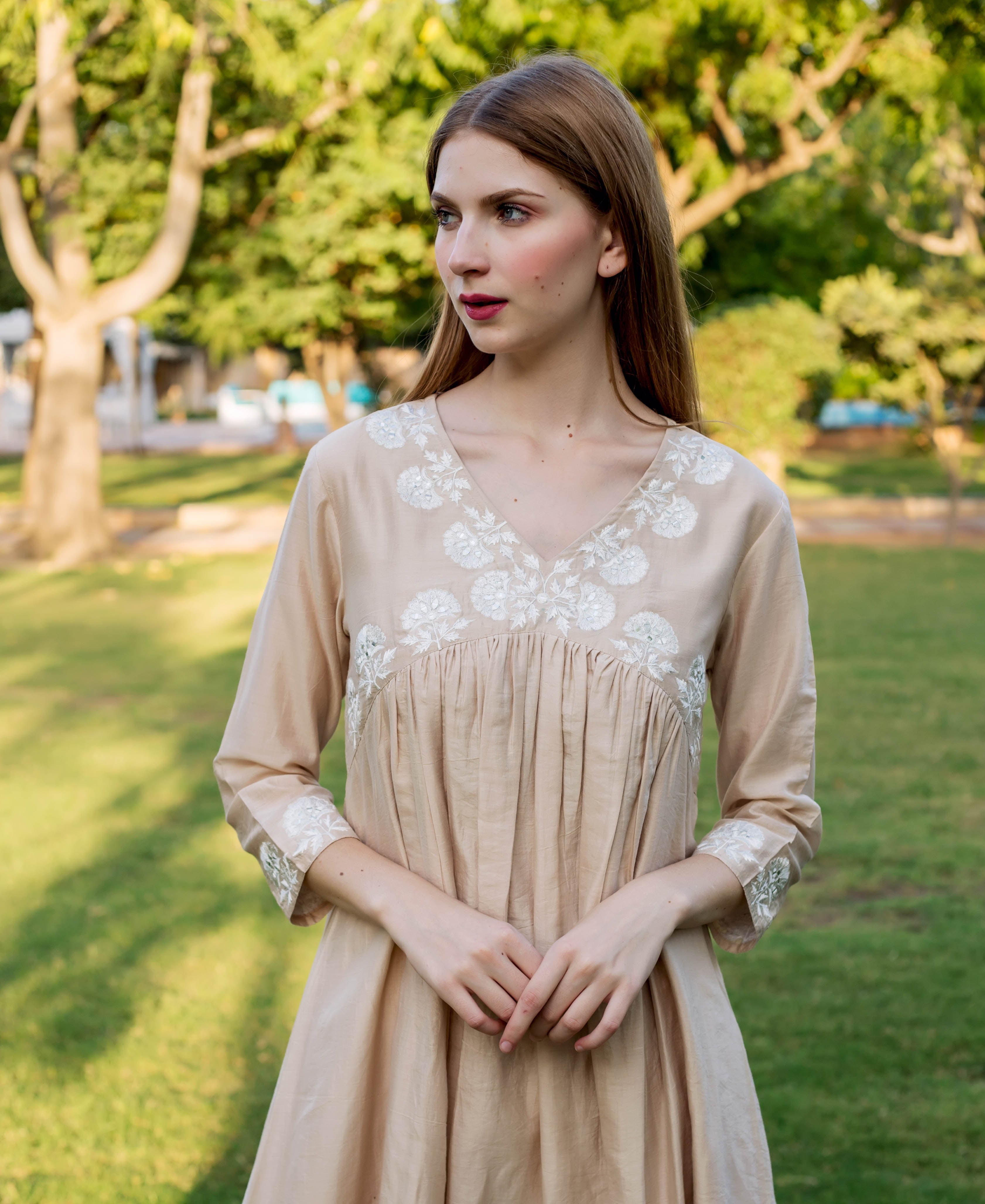 Beige dress with white embroidered floral details on bodice and sleeves, on a female model outdoors.