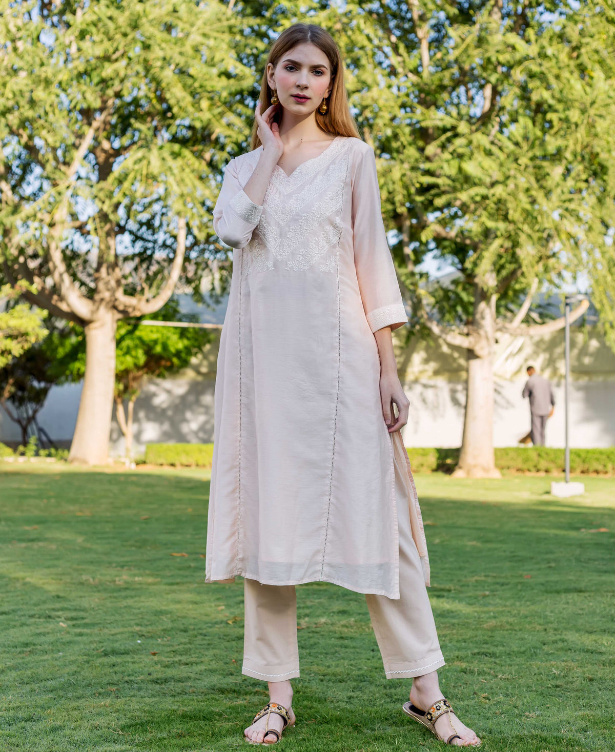 Model wearing a light pink embroidered kurta, matching trousers, and flat sandals outdoors.