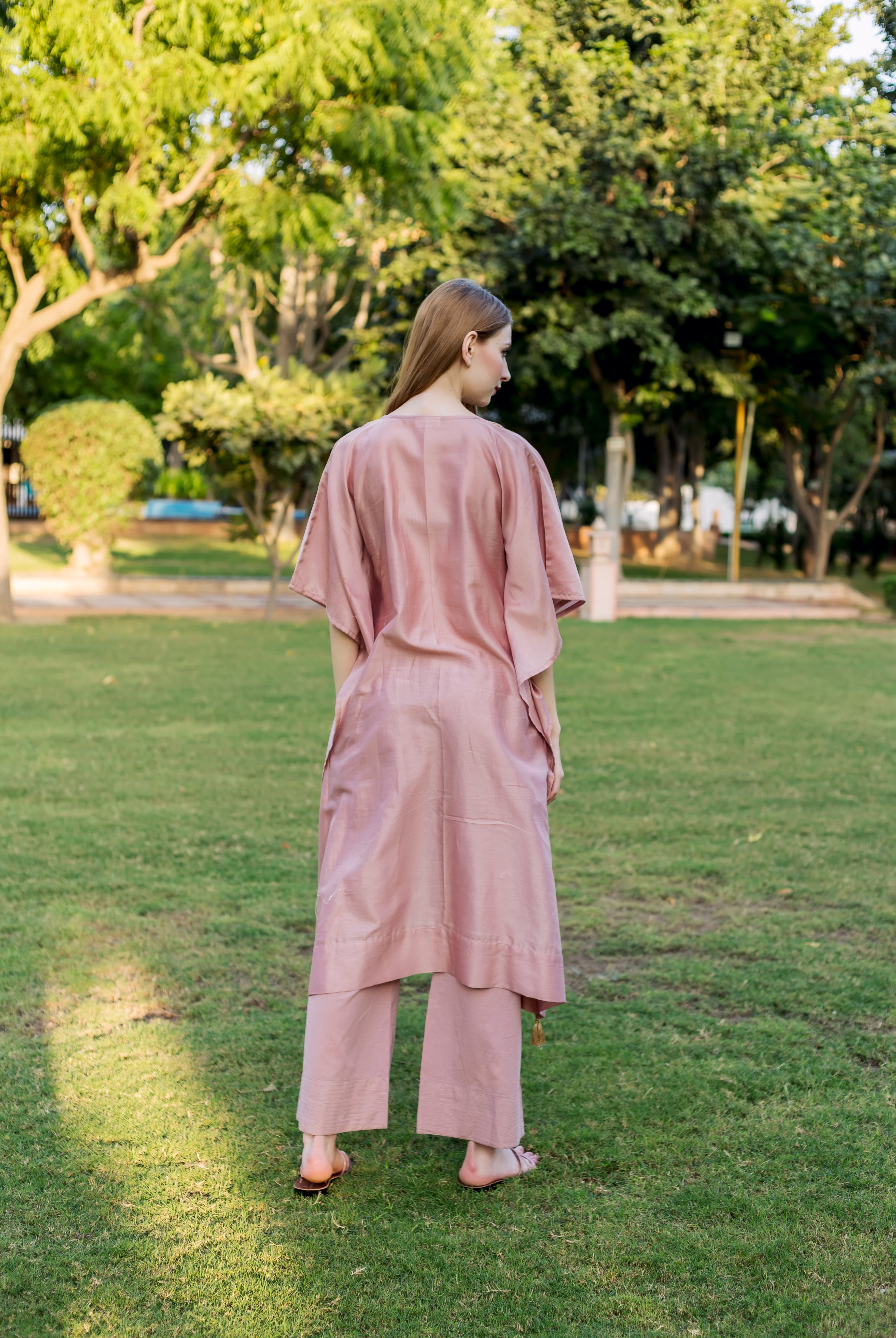 Model in loose-fitting dusty pink kaftan and matching pants, seen from behind in a park.