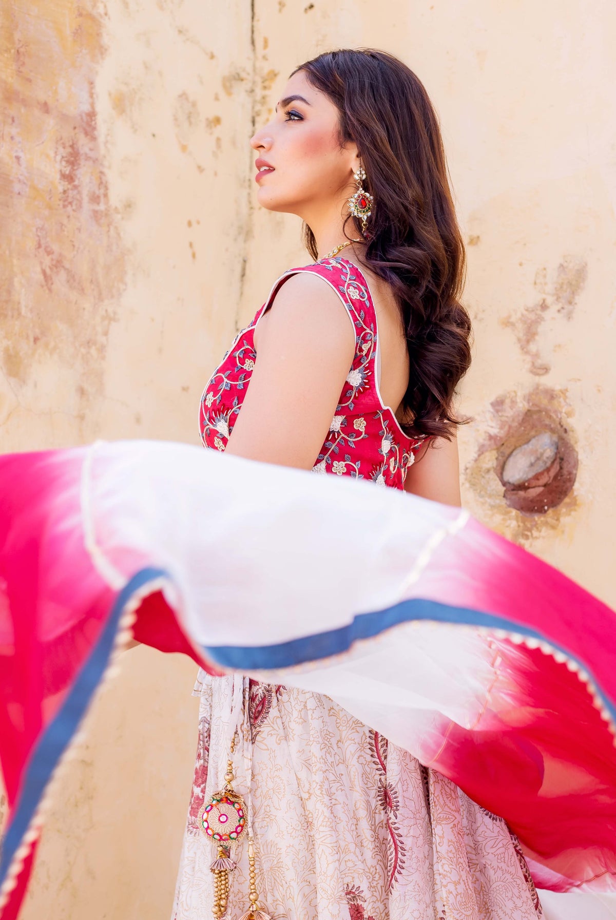 Side view of model in red embroidered top, cream printed lehenga, and flowing pink-white dupatta.