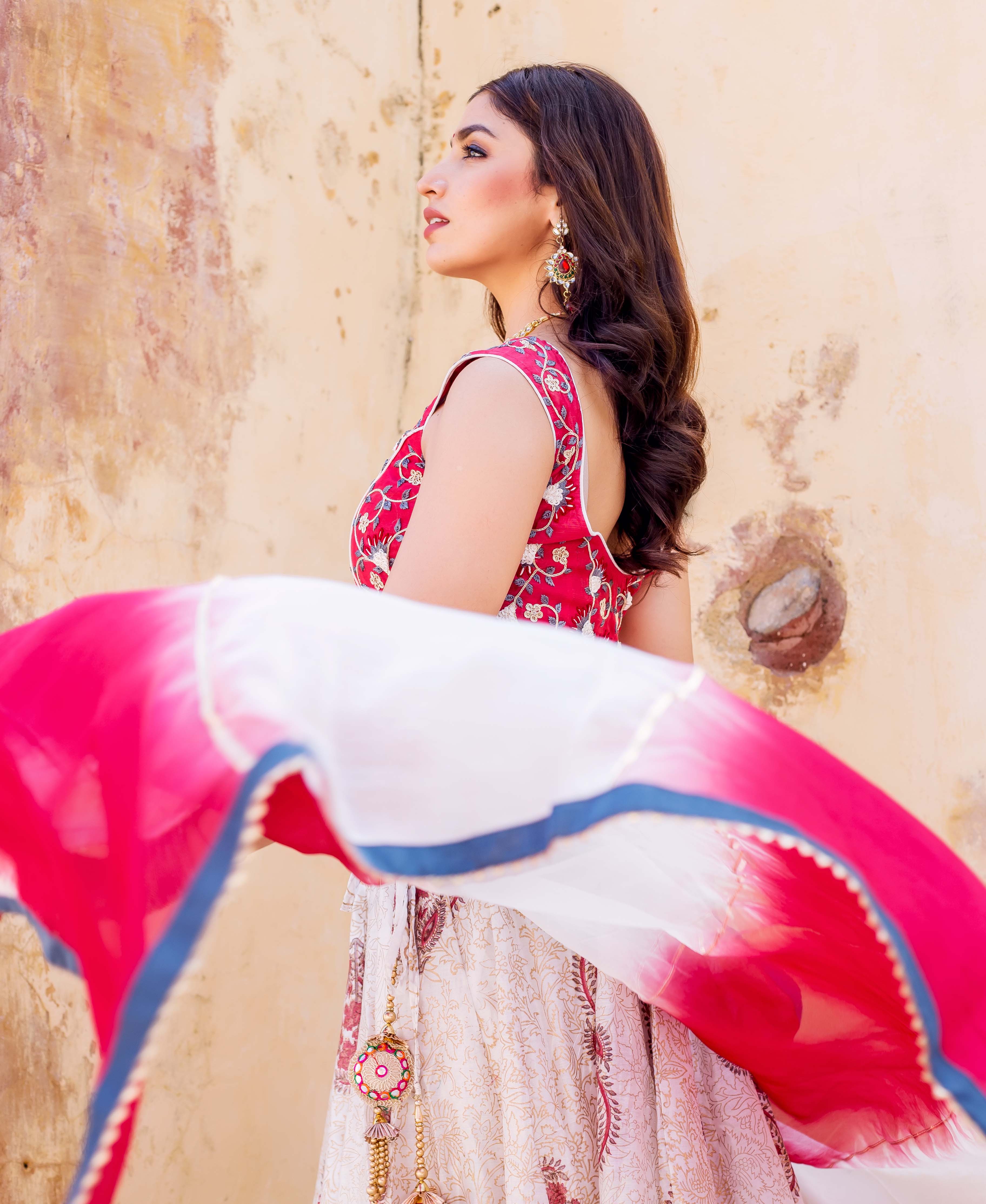 Side view of model in red embroidered top, cream printed lehenga, and flowing pink-white dupatta.