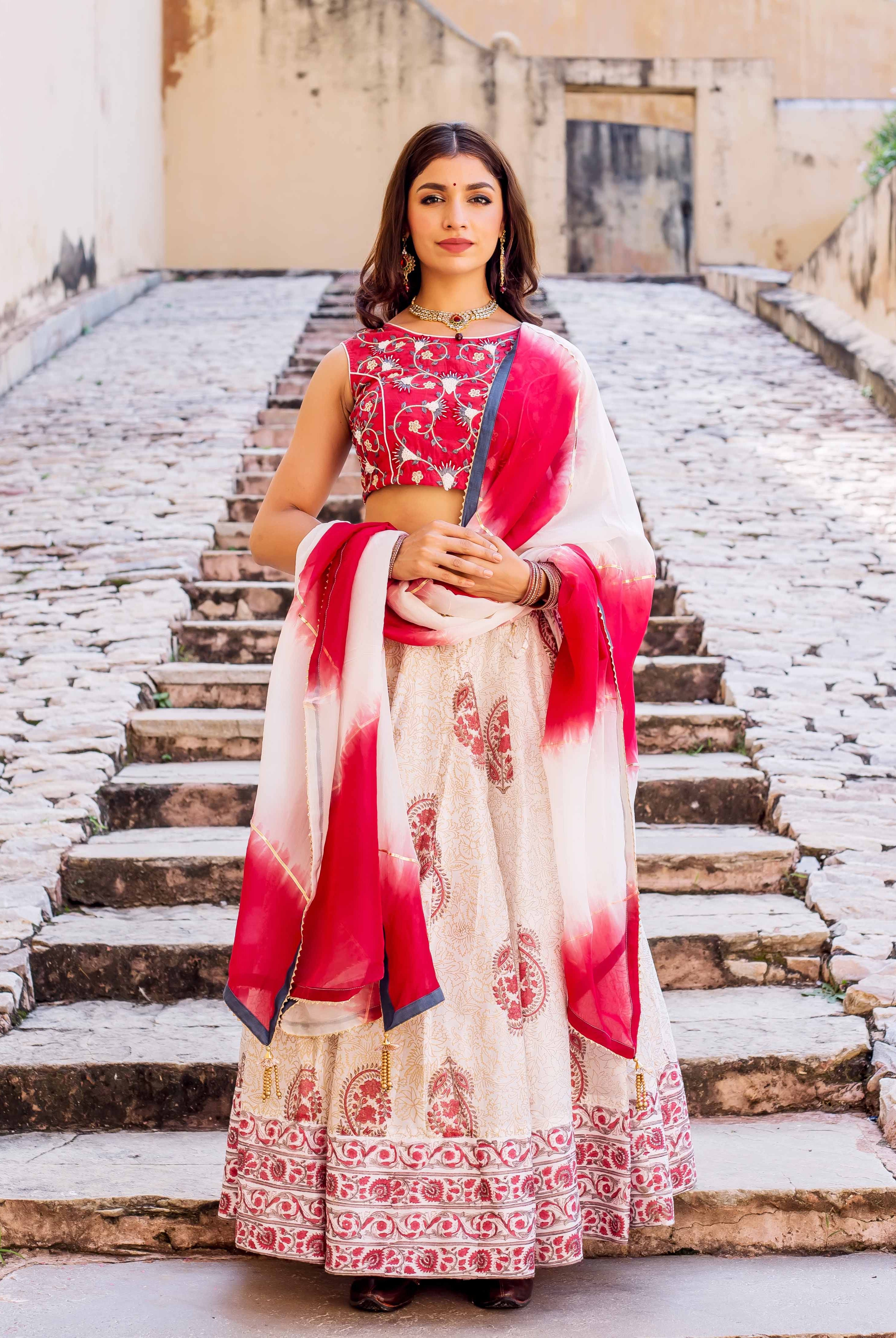 Full-length model wears red embroidered blouse, cream hand block printed lehenga, and red-white dupatta.