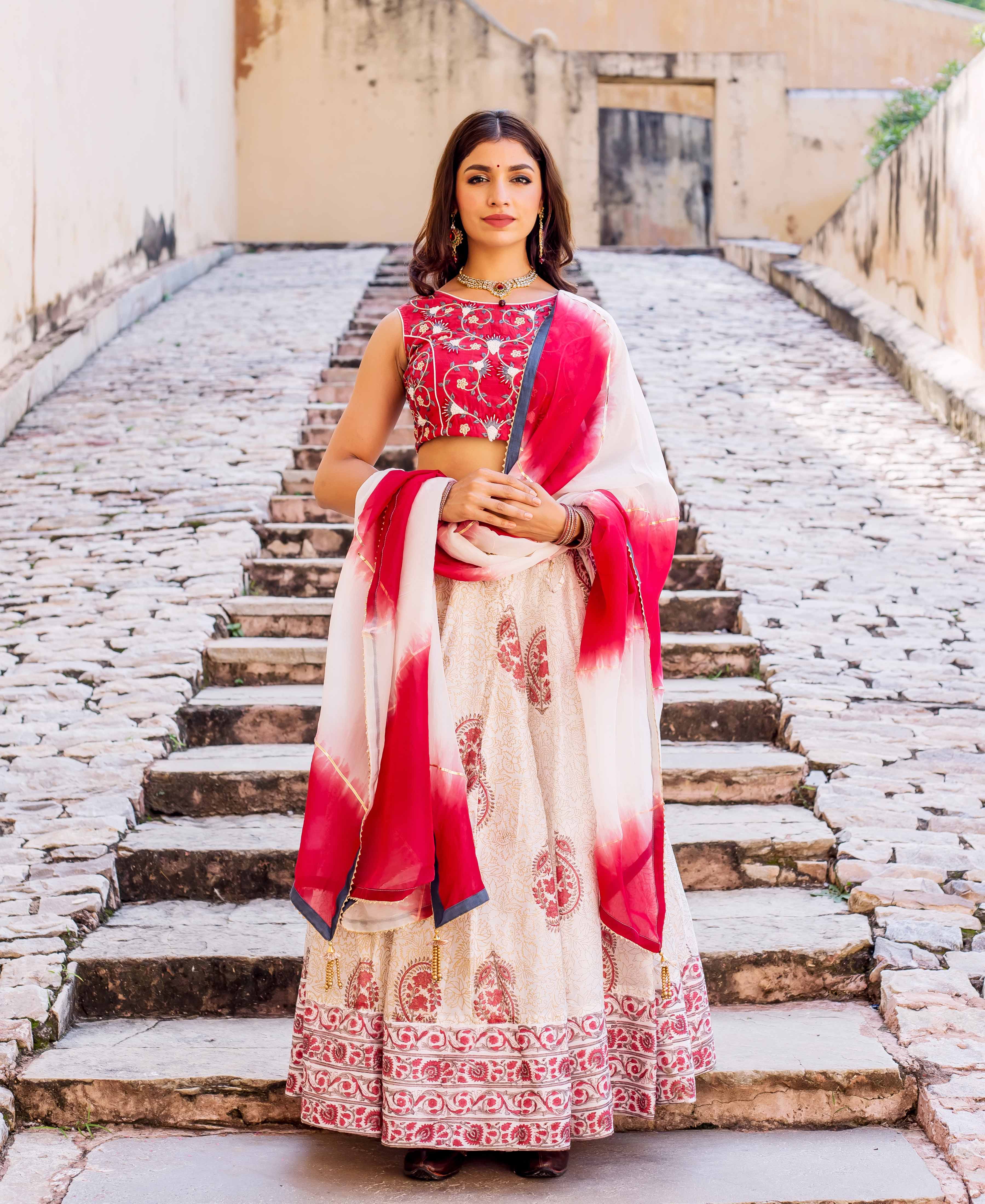 Full-length model wears red embroidered blouse, cream hand block printed lehenga, and red-white dupatta.