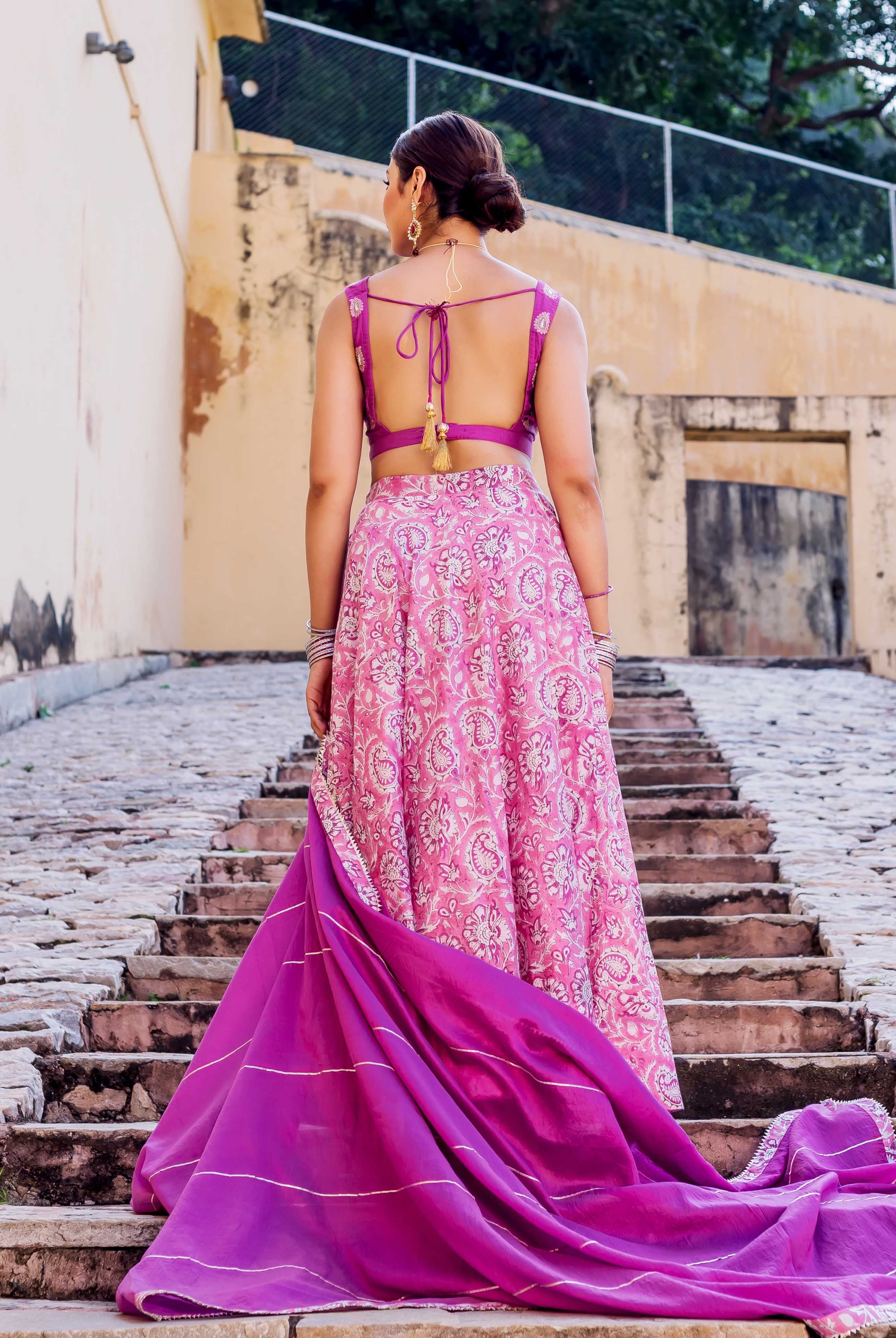 Back view of model in pink hand block printed lehenga, purple blouse, and flowing dupatta.