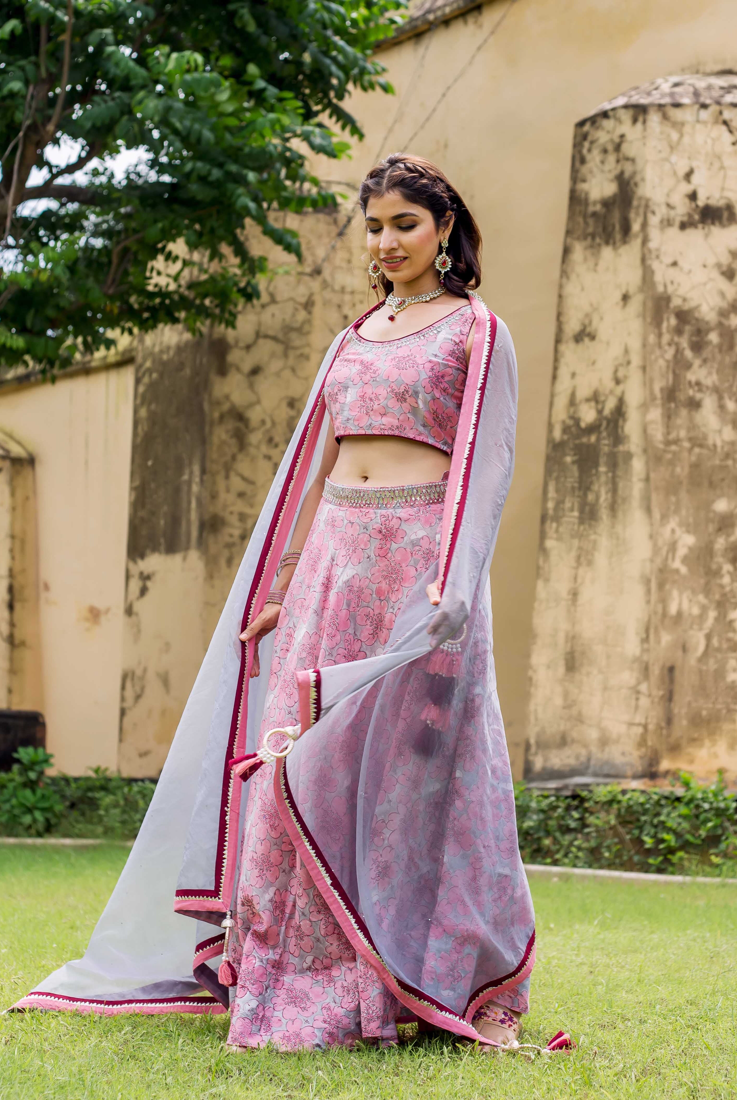 Young woman wearing a pink and grey hand block printed lehenga set, posing in a garden.
