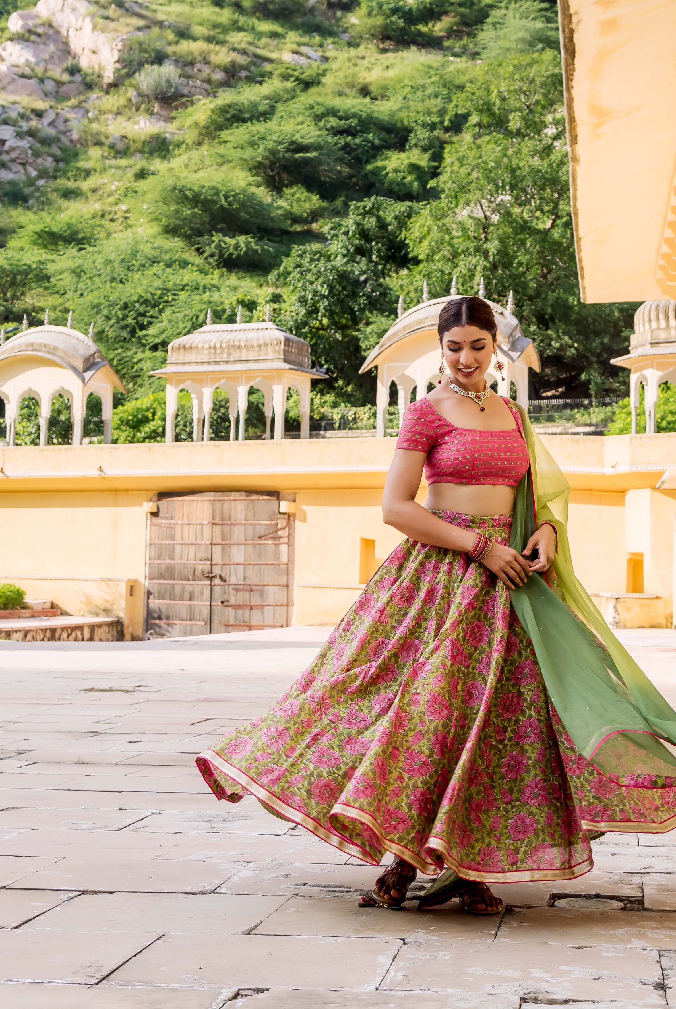 A woman in a pink and green floral lehenga set, posing in front of a building.
