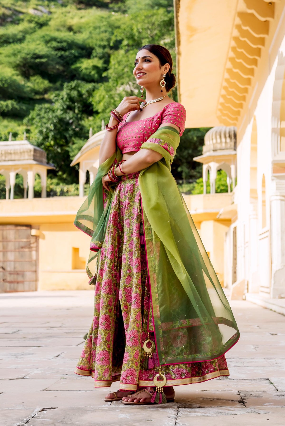 A model wearing a vibrant pink and green lehenga set with intricate floral patterns.