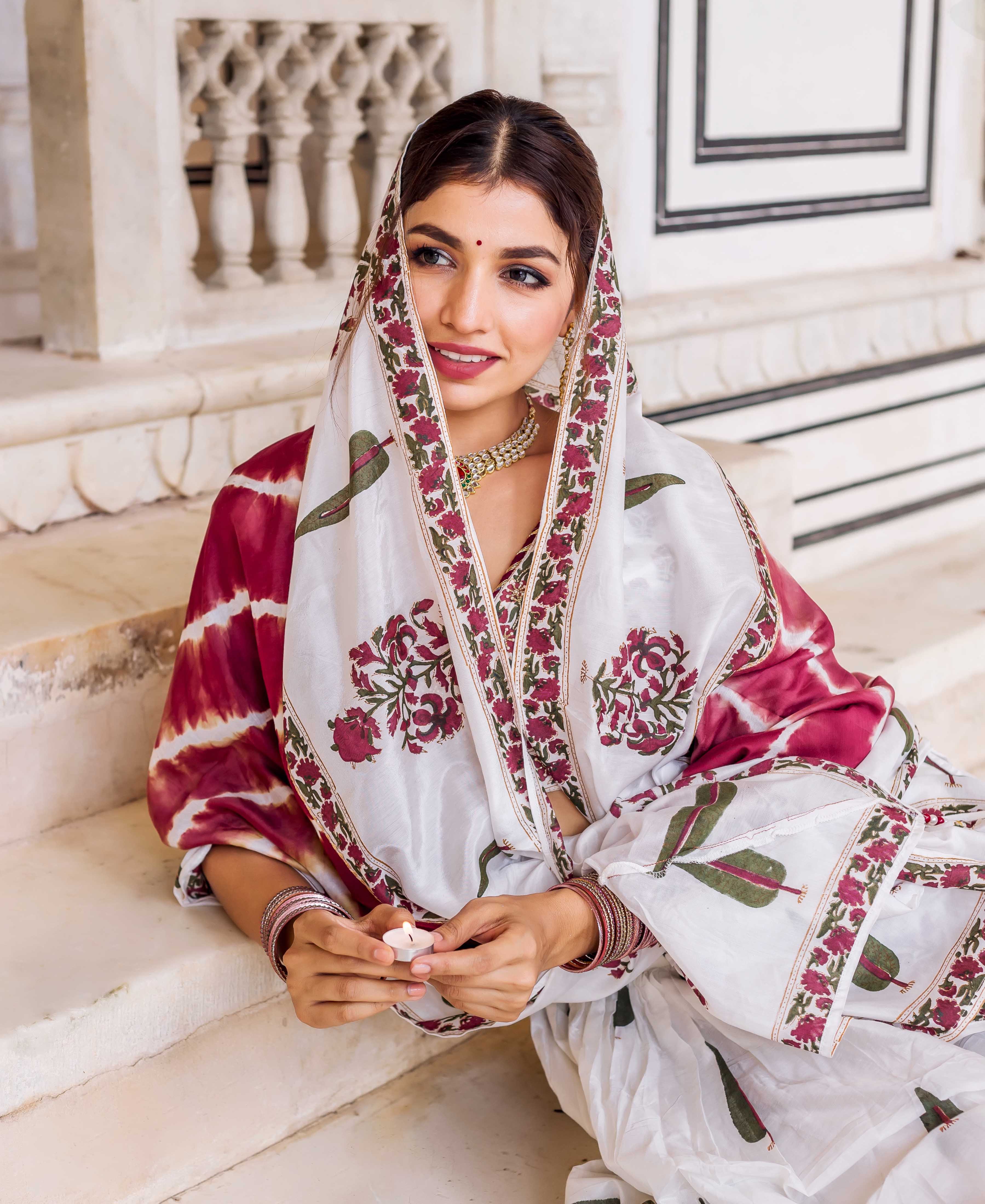 Model seated, wearing a white and maroon hand-block printed lehenga set with floral and tie-dye patterns.