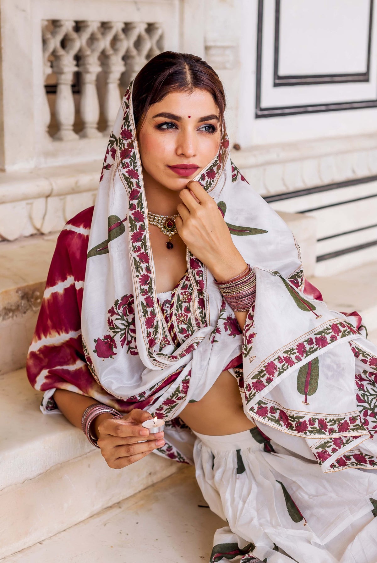 Seated model in white hand block printed lehenga set with maroon and green floral patterns.