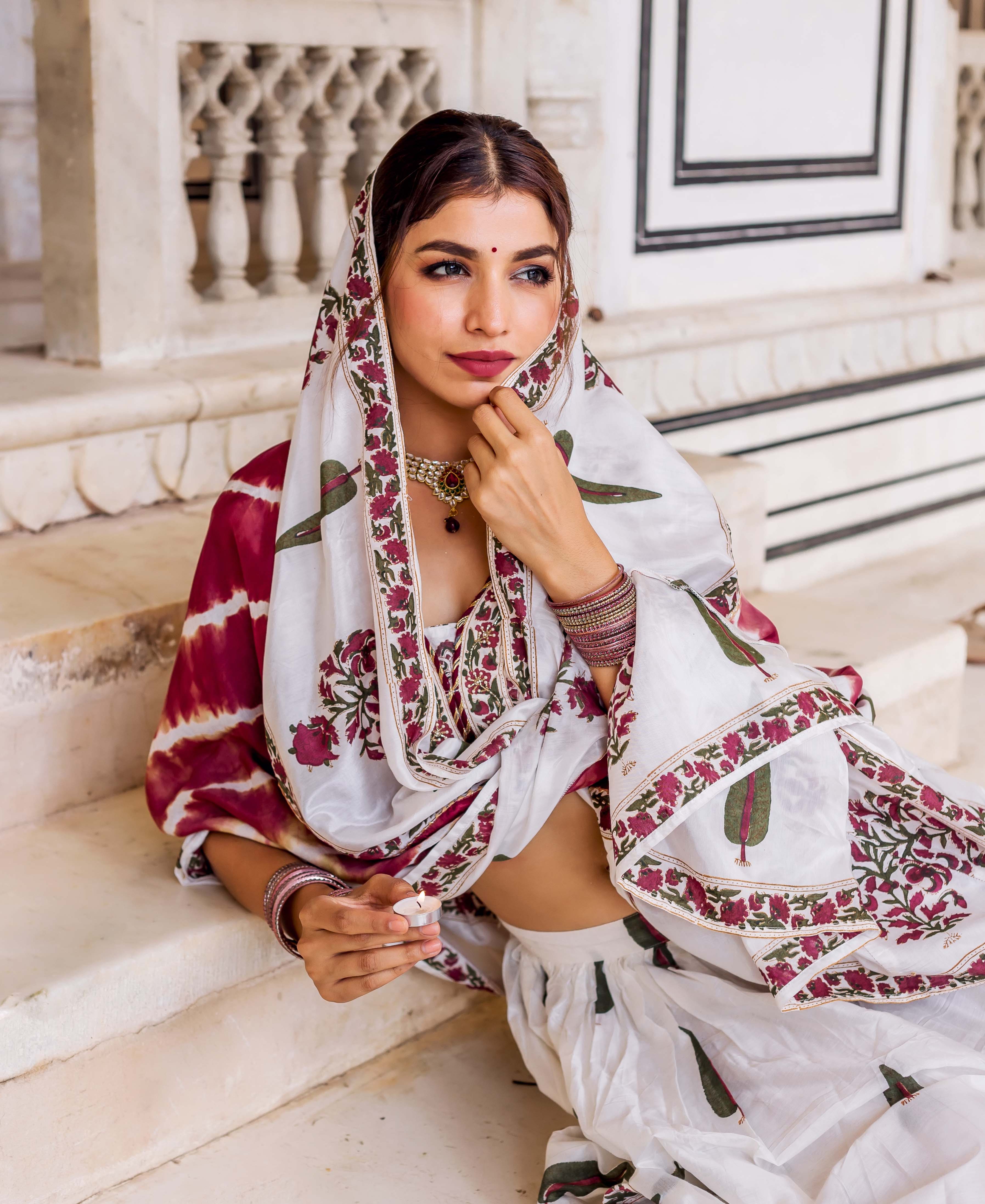 Seated model in white hand block printed lehenga set with maroon and green floral patterns.