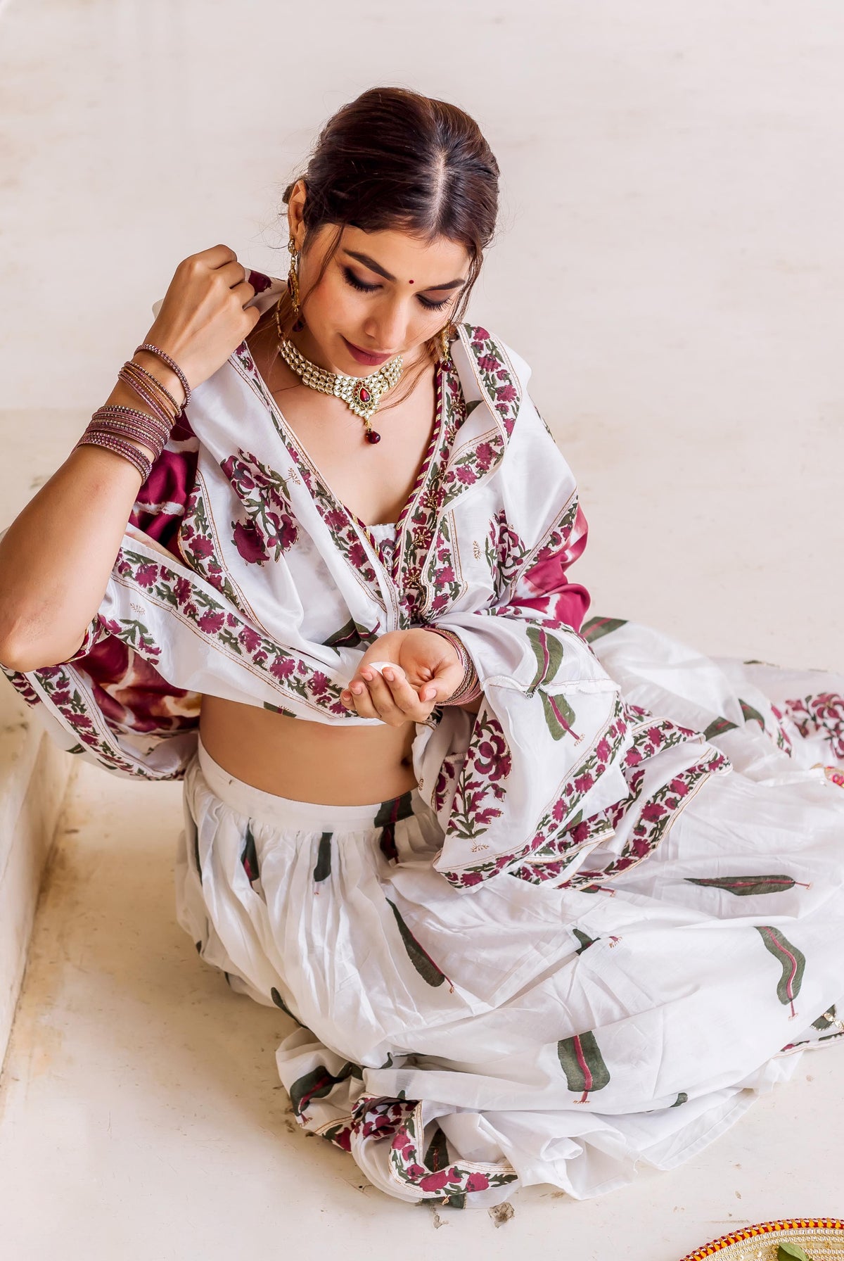 Sitting model in white hand block printed lehenga set with maroon floral and green leaf patterns.