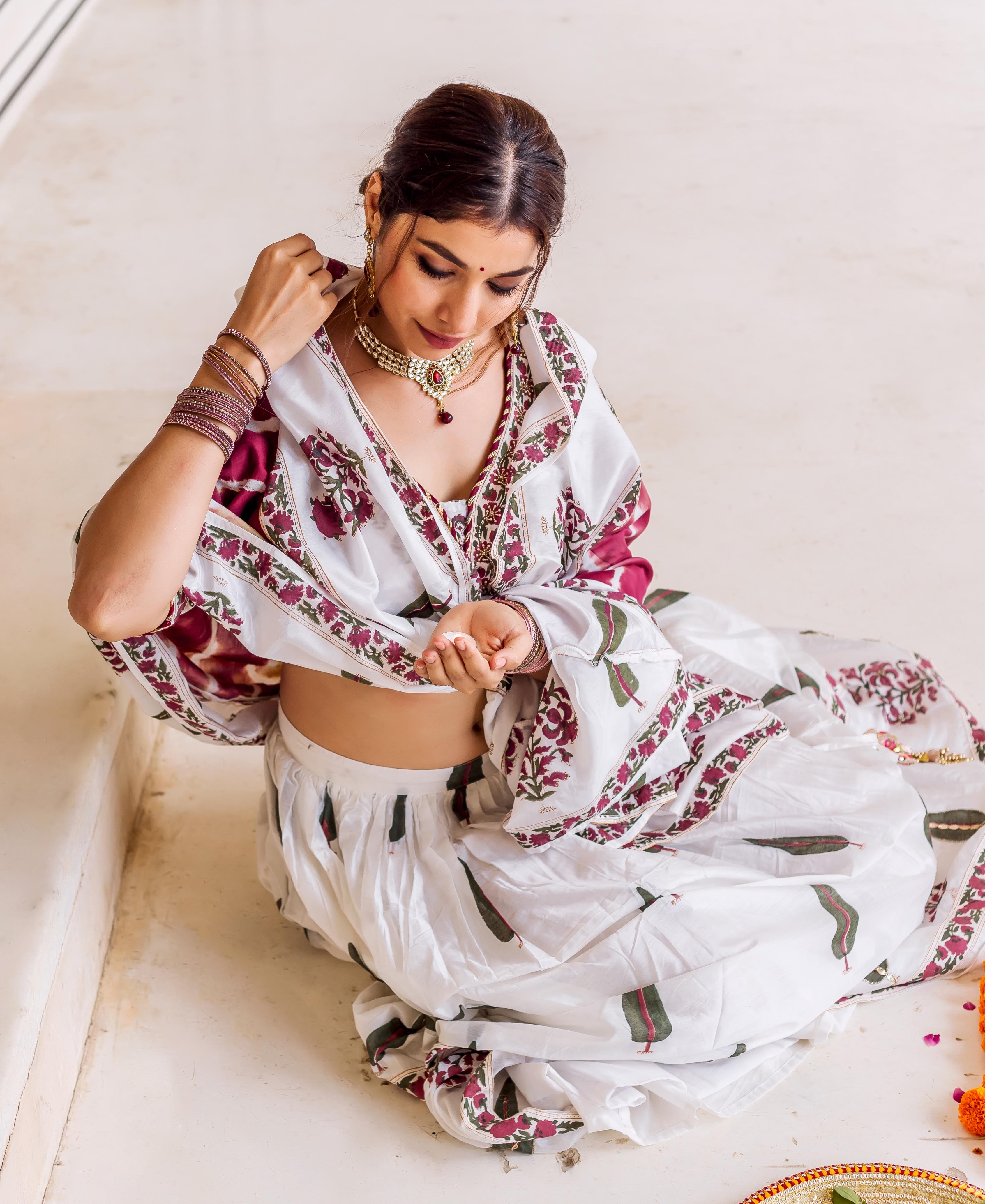 Sitting model in white hand block printed lehenga set with maroon floral and green leaf patterns.