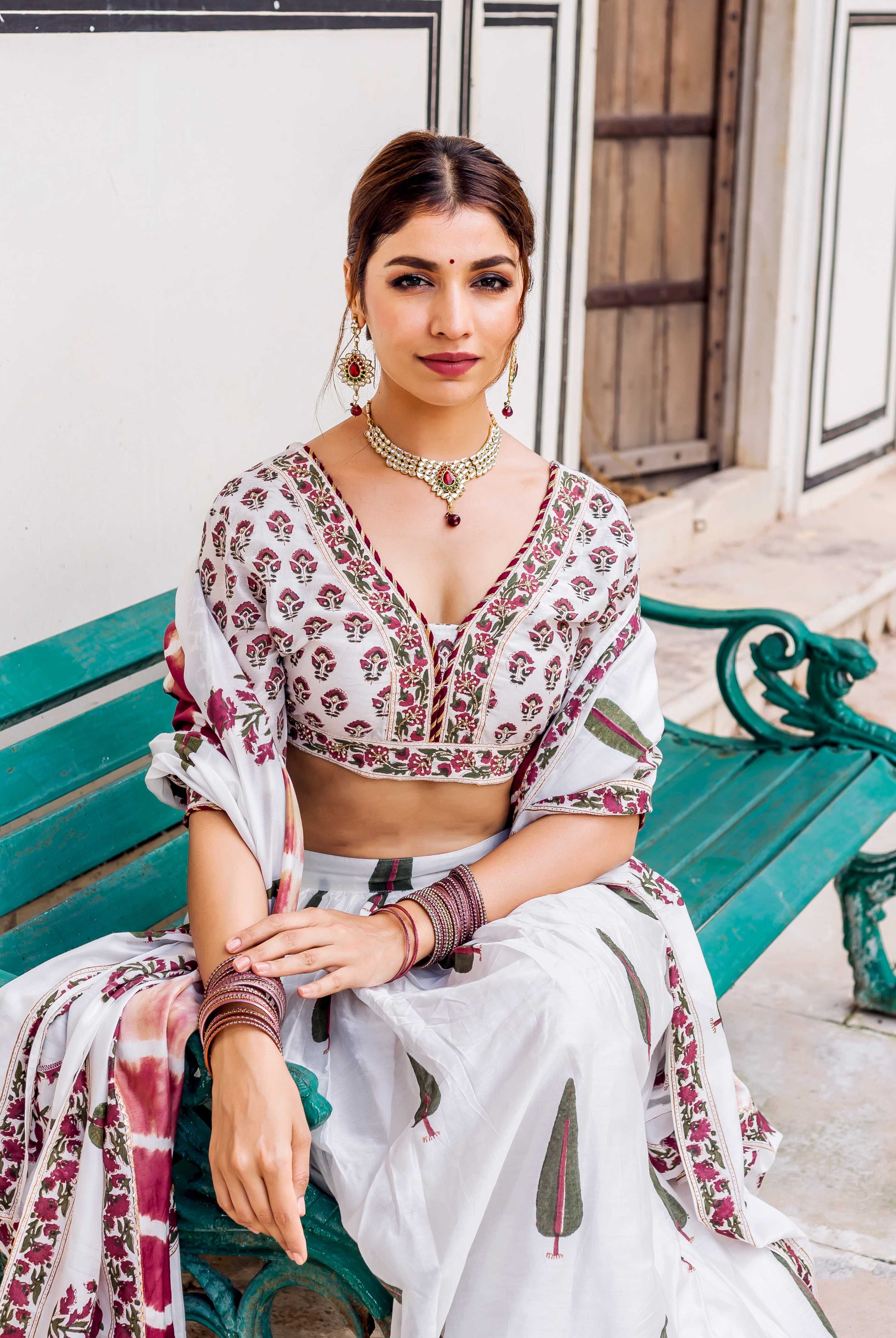 Model seated, showcasing white hand-block printed lehenga set with maroon flowers and green trees.
