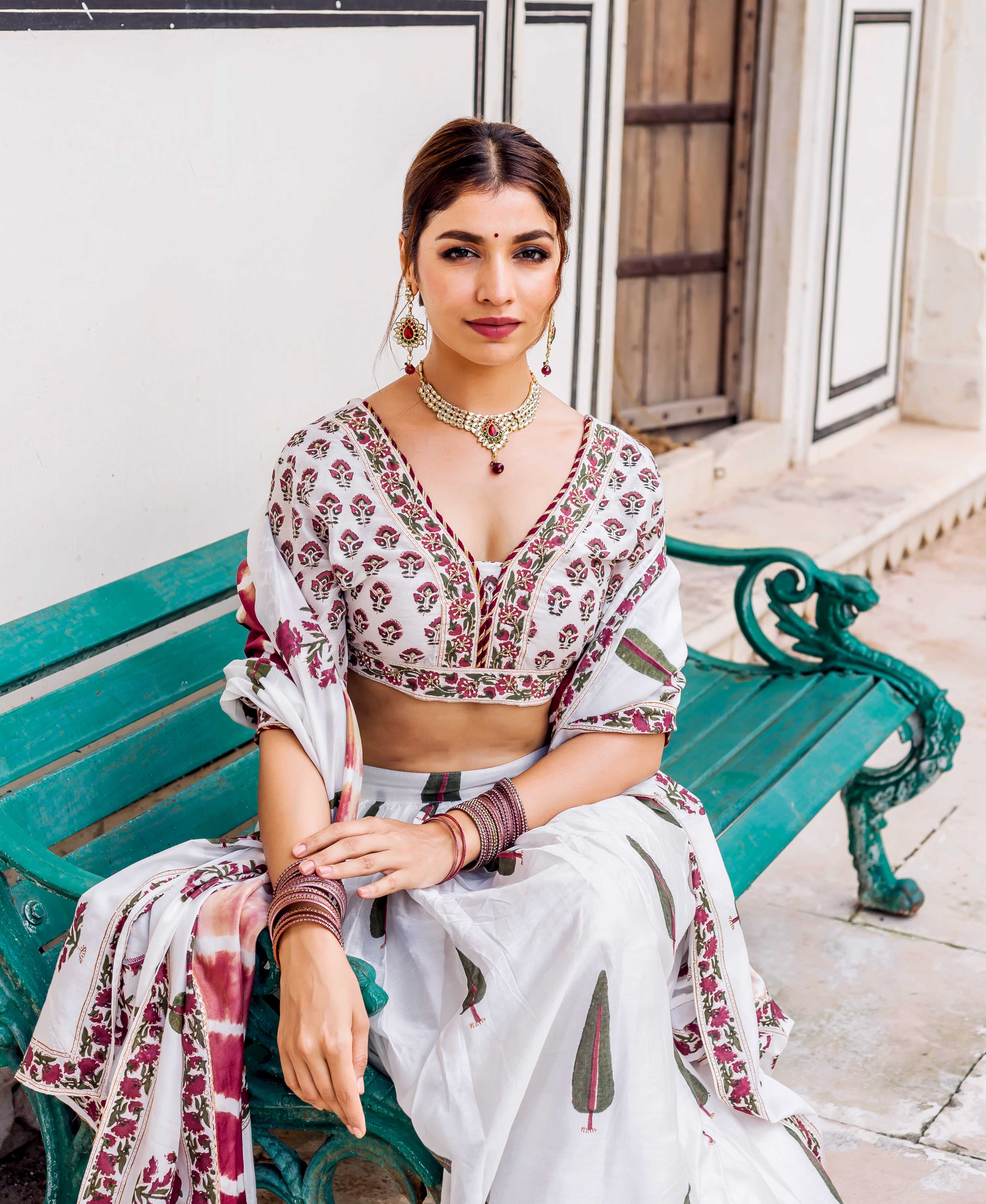 Model seated, showcasing white hand-block printed lehenga set with maroon flowers and green trees.