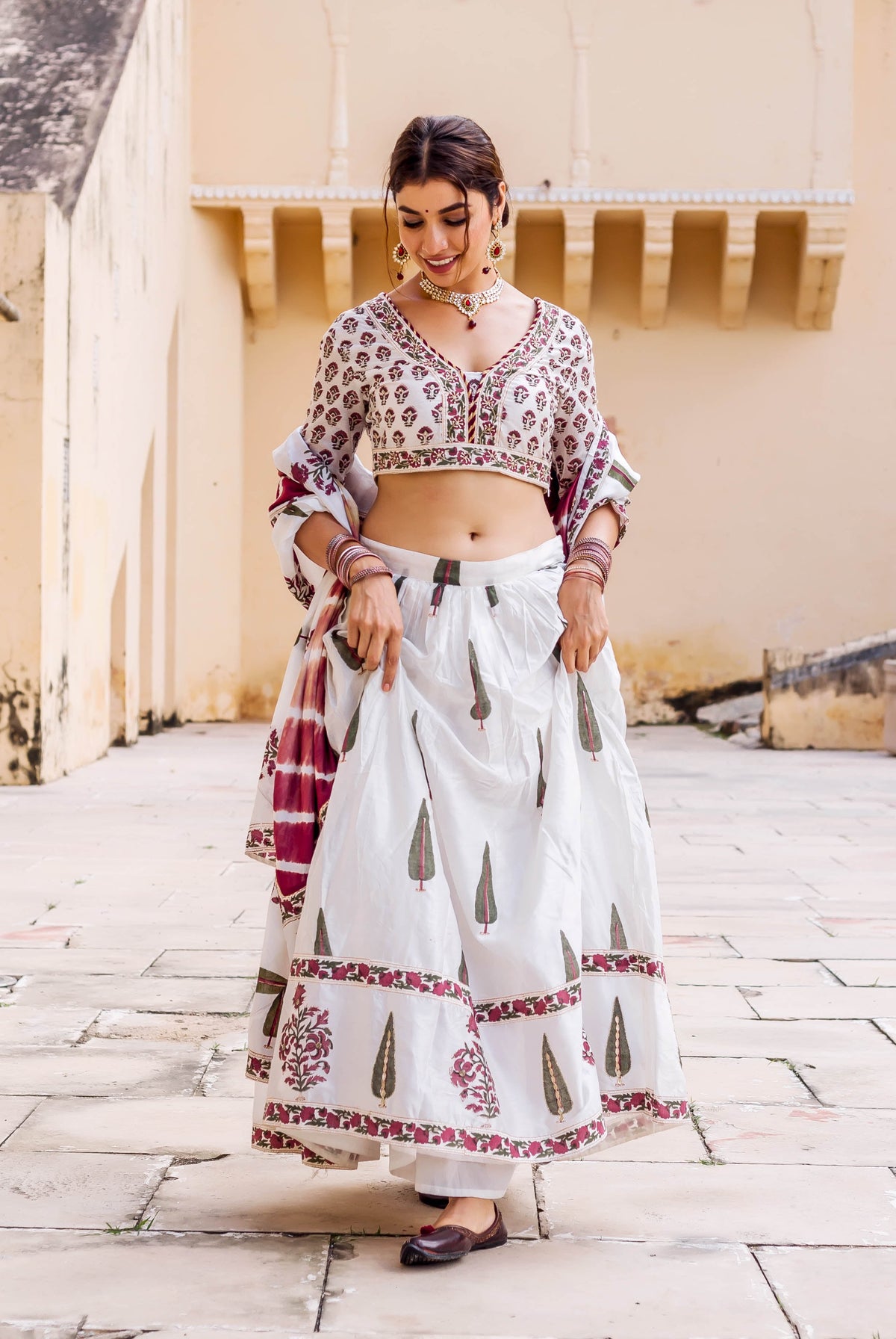 Woman in white hand-block-printed lehenga with maroon and green accents, smiling downward.