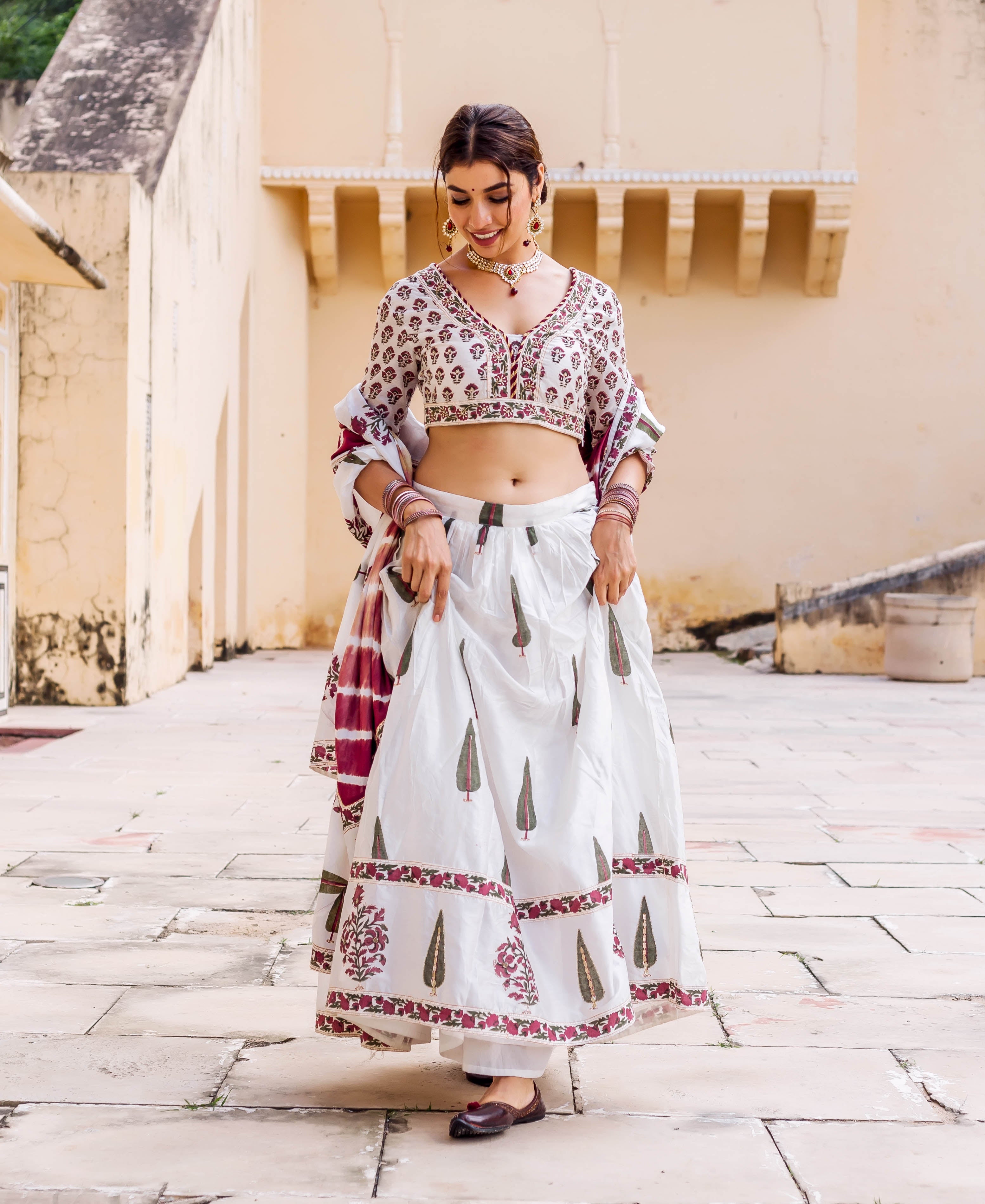 Woman in white hand-block-printed lehenga with maroon and green accents, smiling downward.