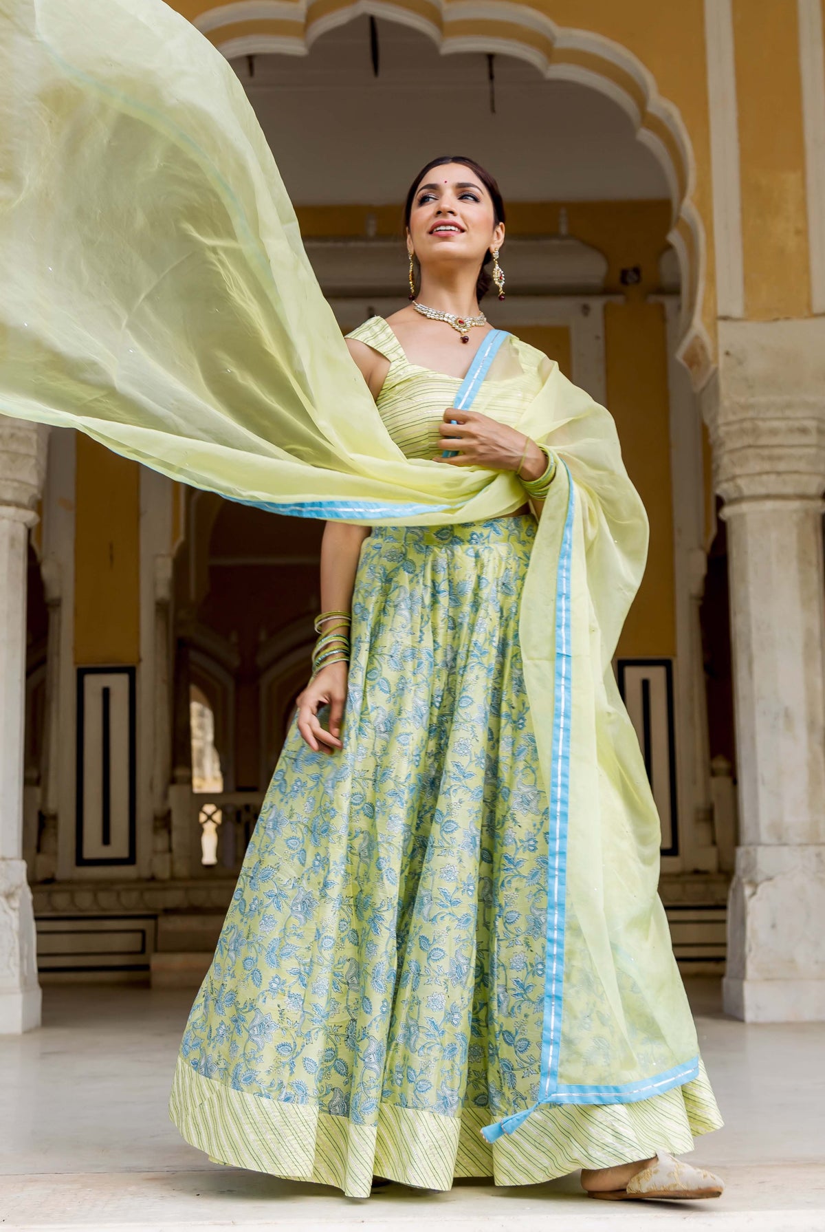 Model wearing lime green hand-block printed lehenga set with blue floral patterns and flowing dupatta.