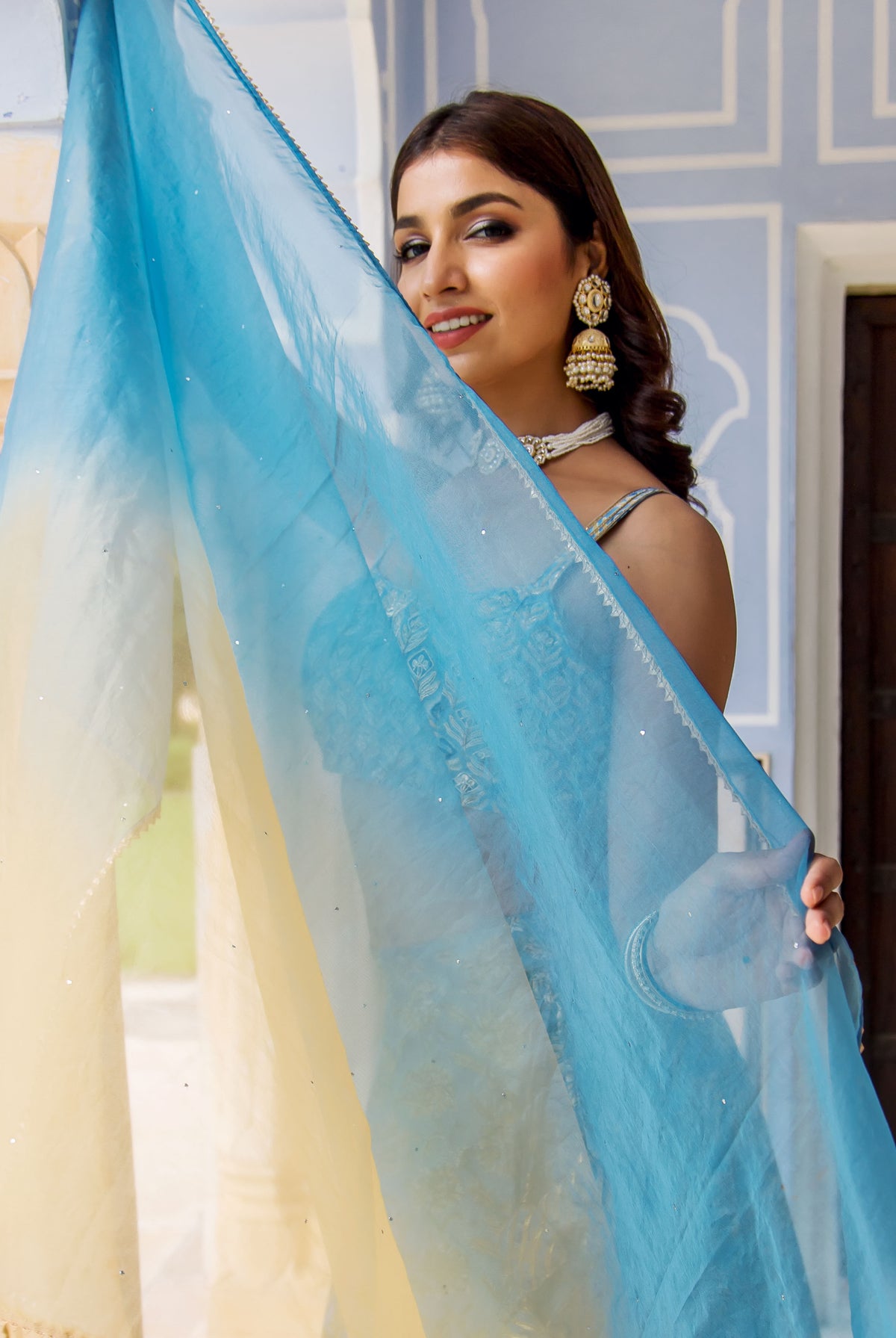 Model in a white lehenga set peeking behind an embroidered ombre yellow-blue dupatta.