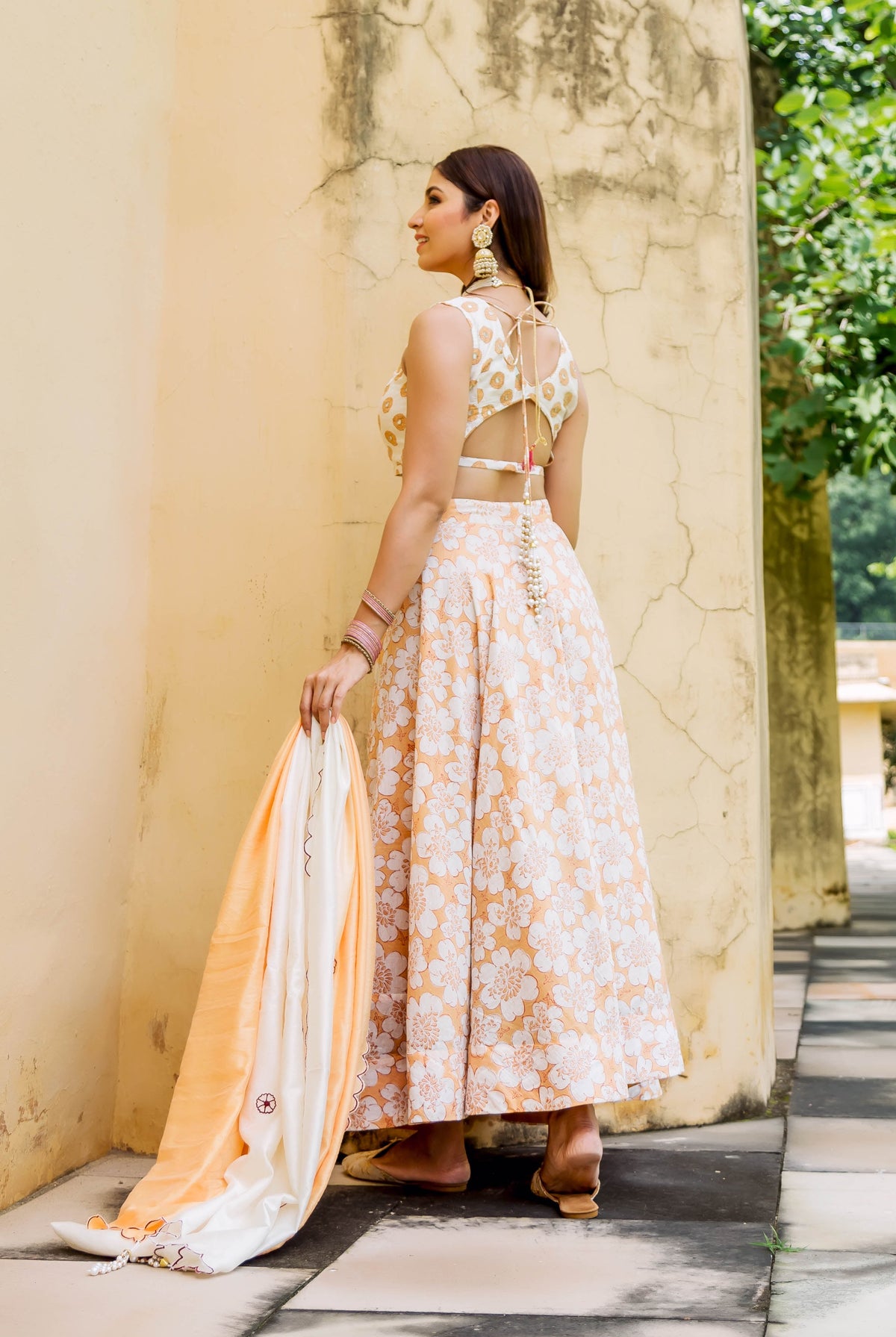 Woman in orange and white floral lehenga set with embroidery, standing outdoors, back to camera.