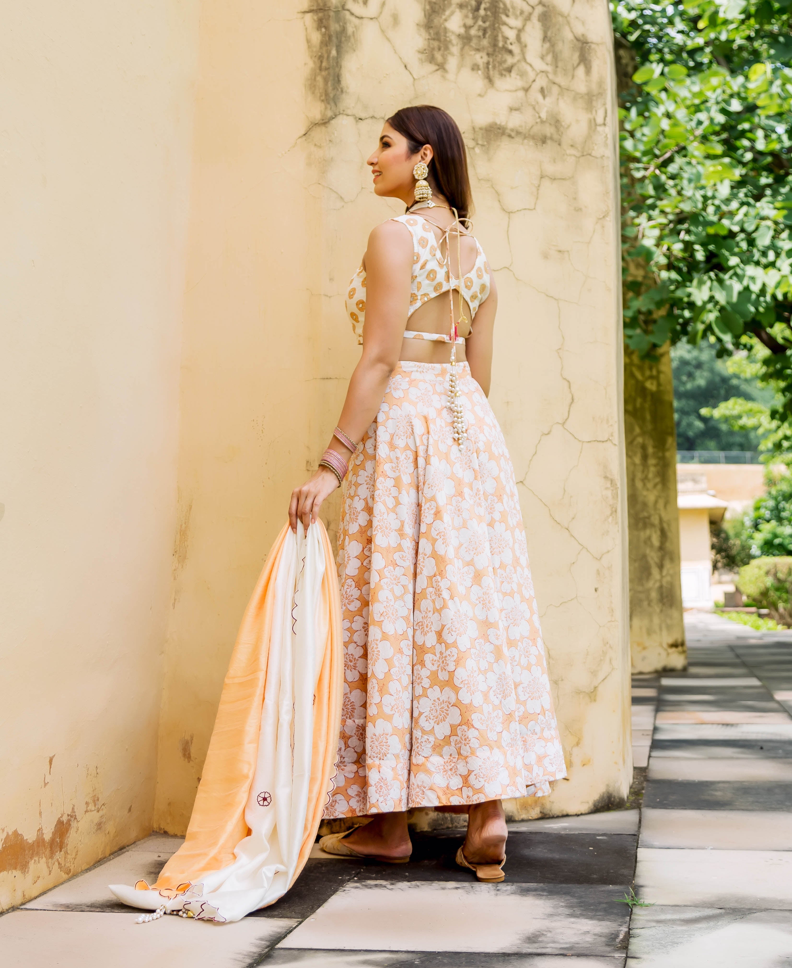 Woman in orange and white floral lehenga set with embroidery, standing outdoors, back to camera.