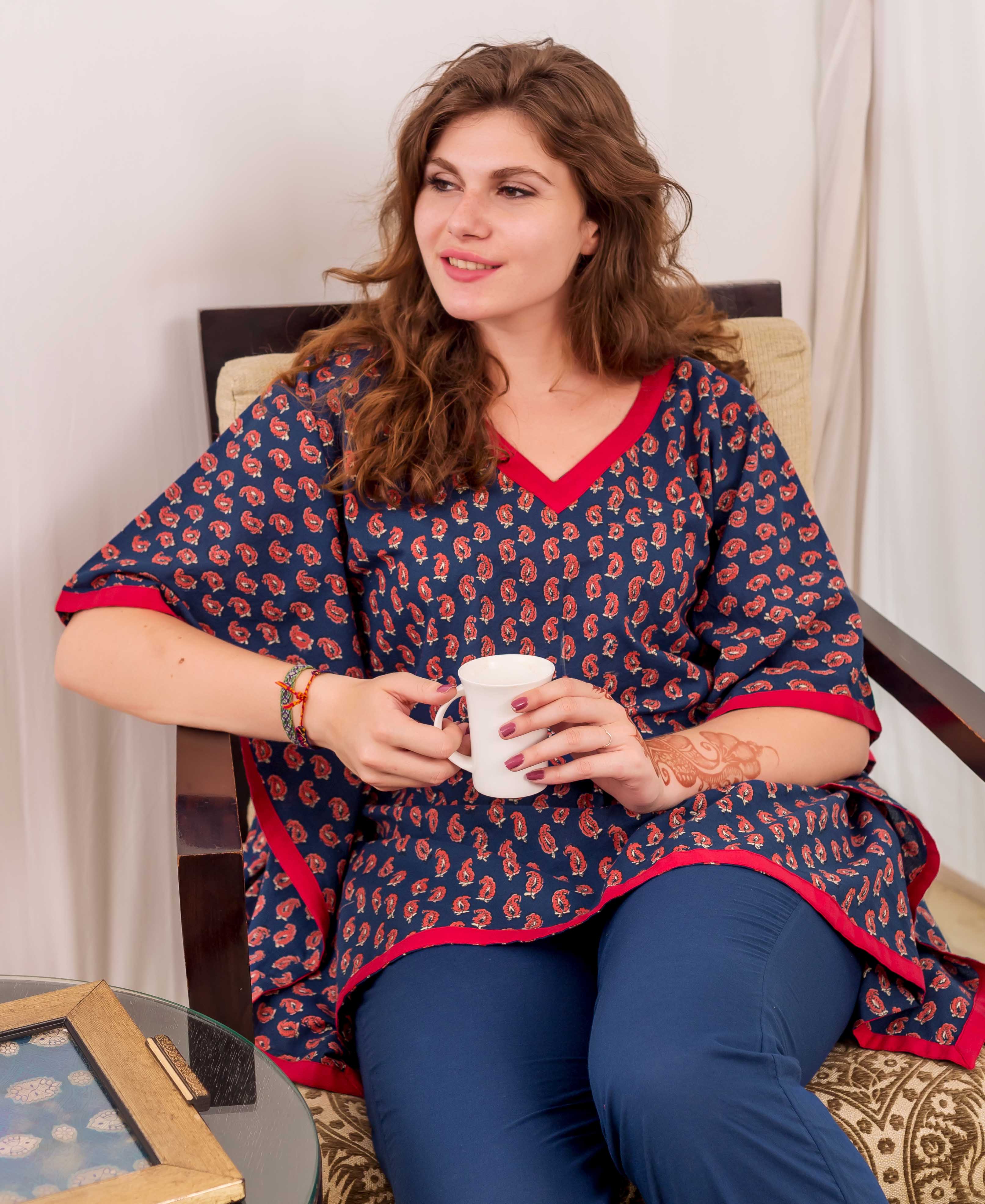 A woman sits in a chair, wearing a blue and red kaftan top with a floral print.