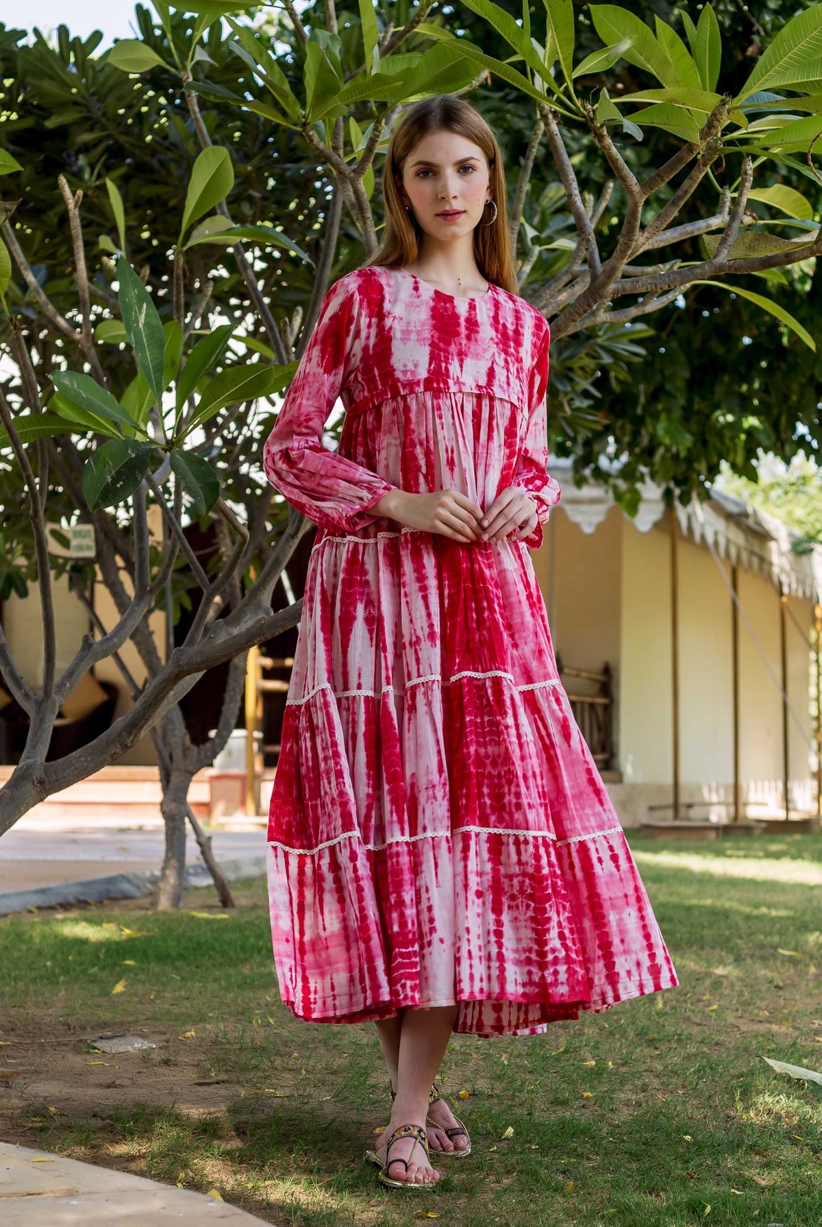 Woman in a vibrant red tie-dye tiered dress with long sleeves, standing in a garden.