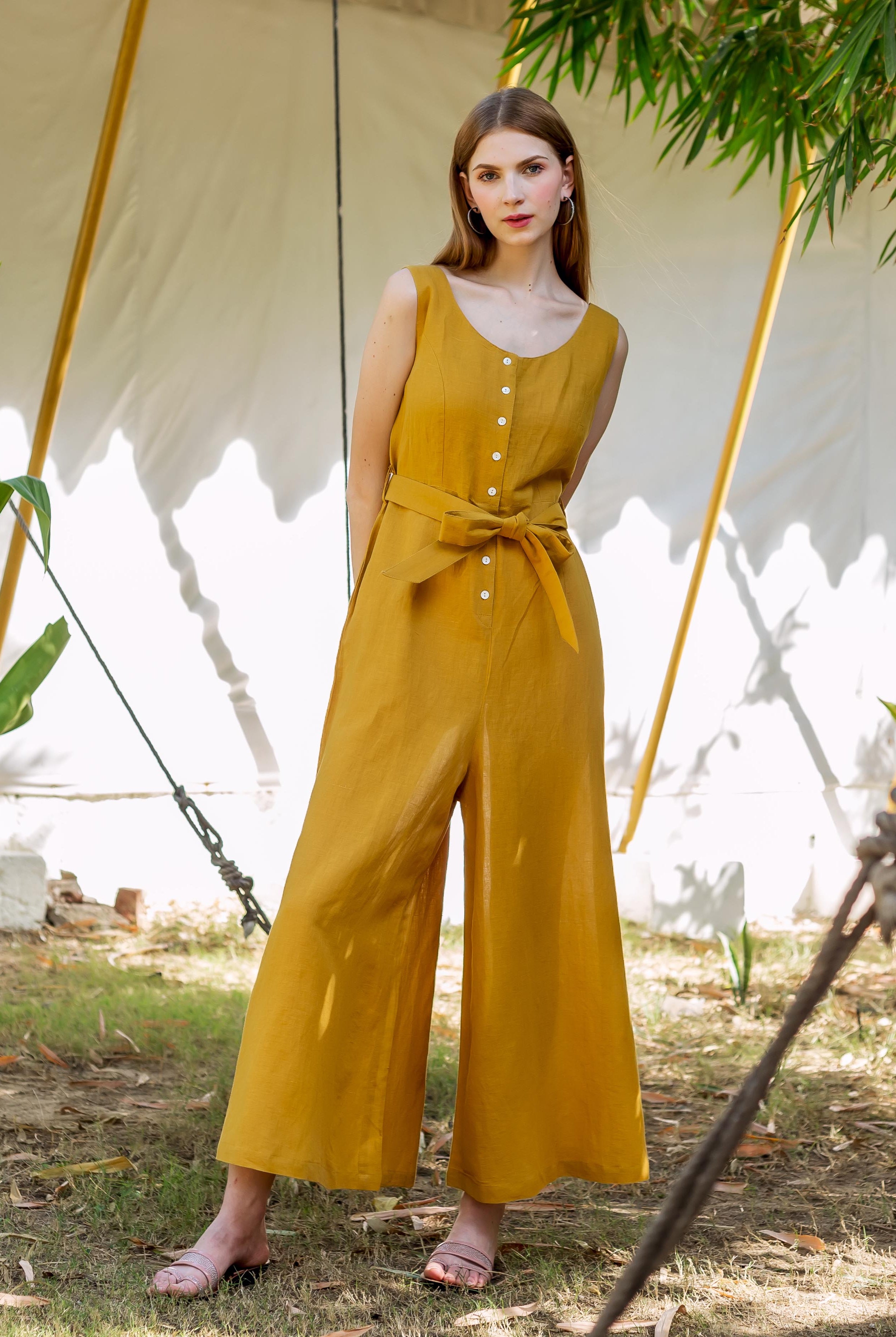 Model stands in full shot, wearing an ochre sleeveless wide-leg jumpsuit with tie belt.
