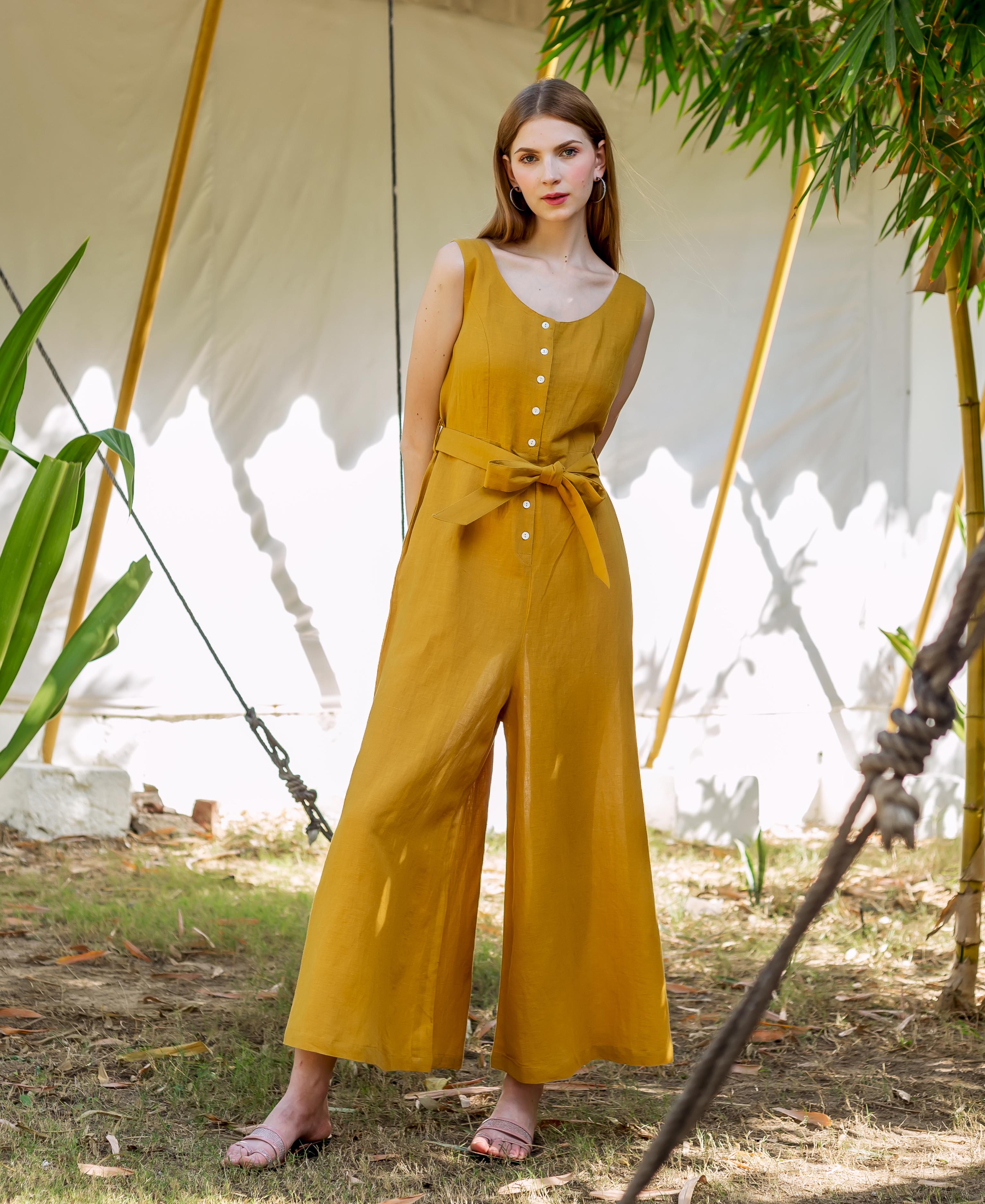 Model stands in full shot, wearing an ochre sleeveless wide-leg jumpsuit with tie belt.