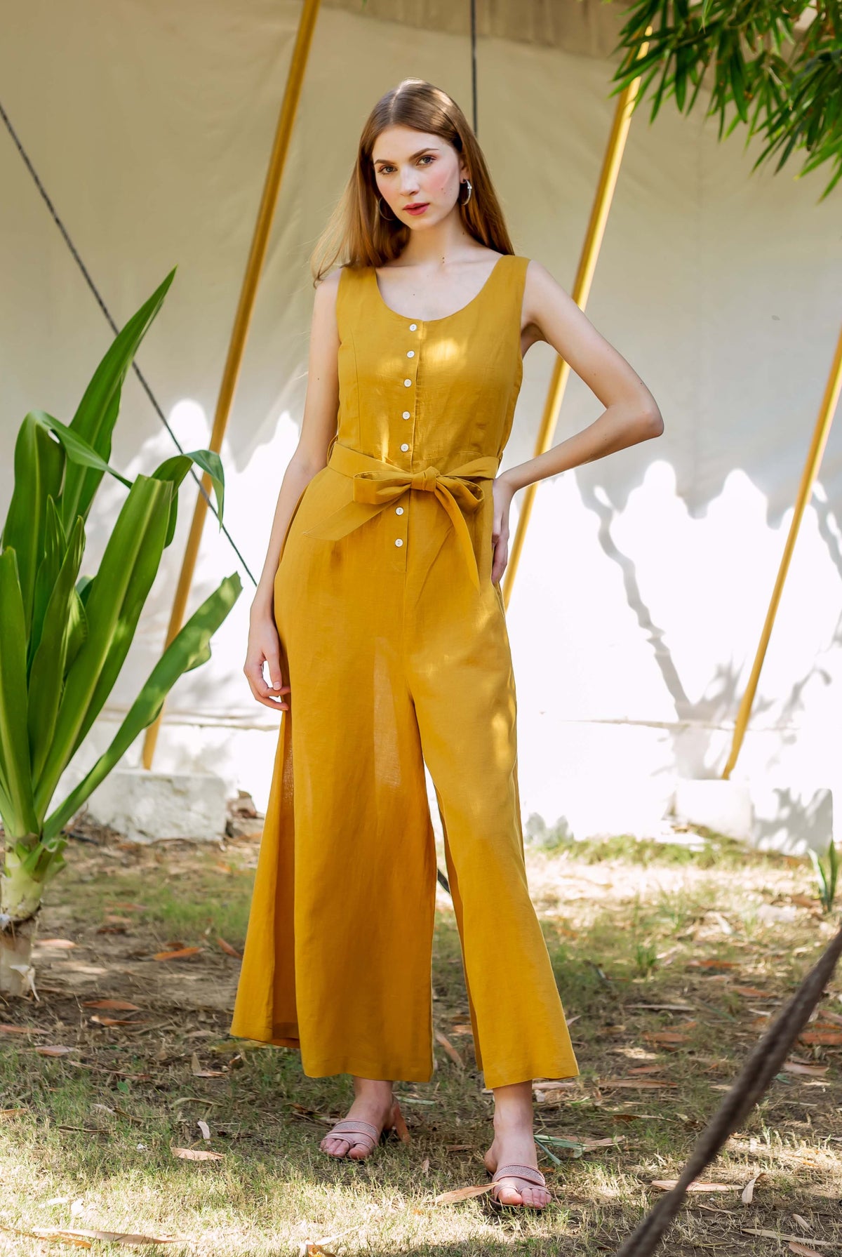 Woman in an ochre sleeveless jumpsuit with button front, tie-waist, and wide legs.
