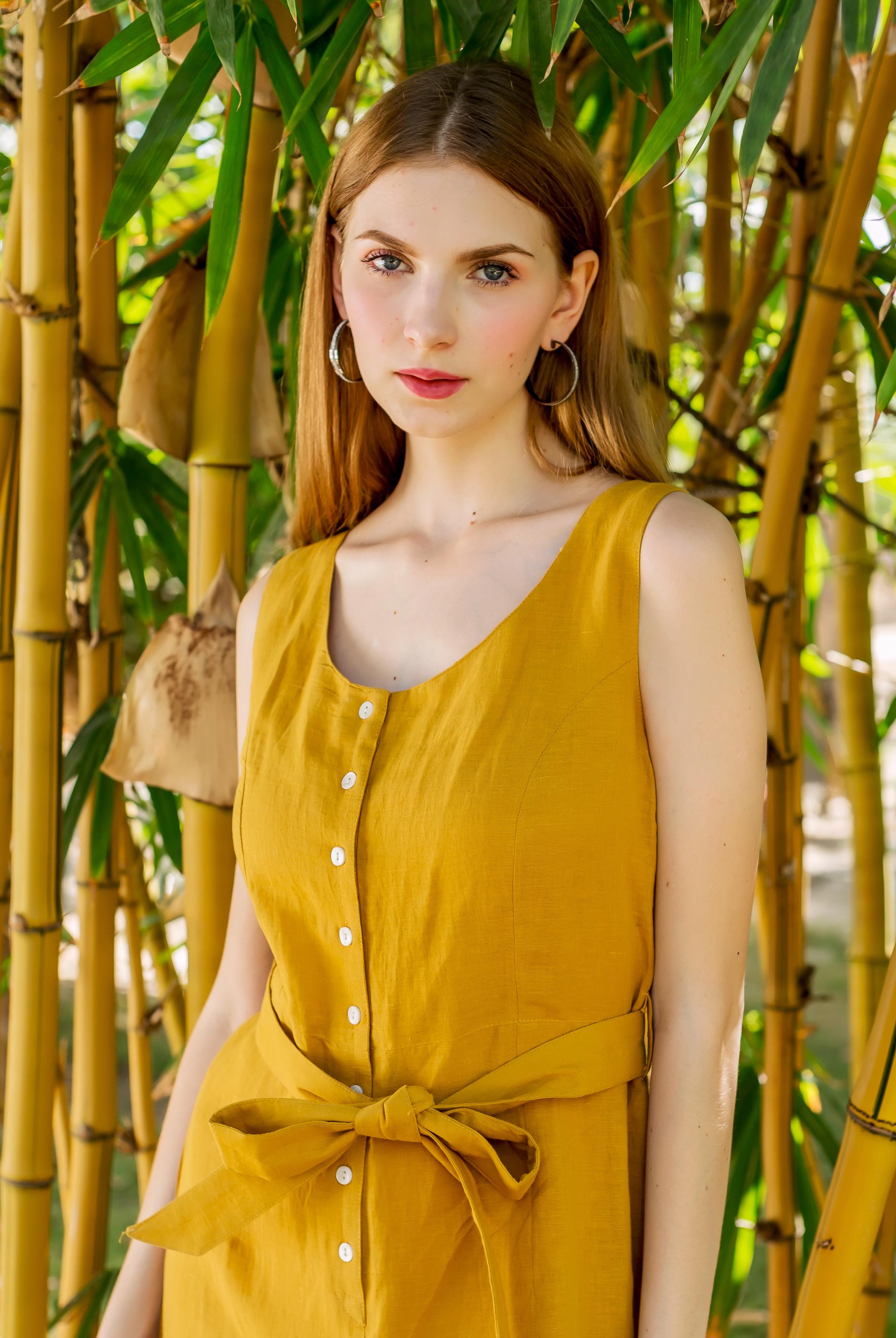 Half-length shot of a model wearing an ochre sleeveless jumpsuit with front buttons and tied waist.