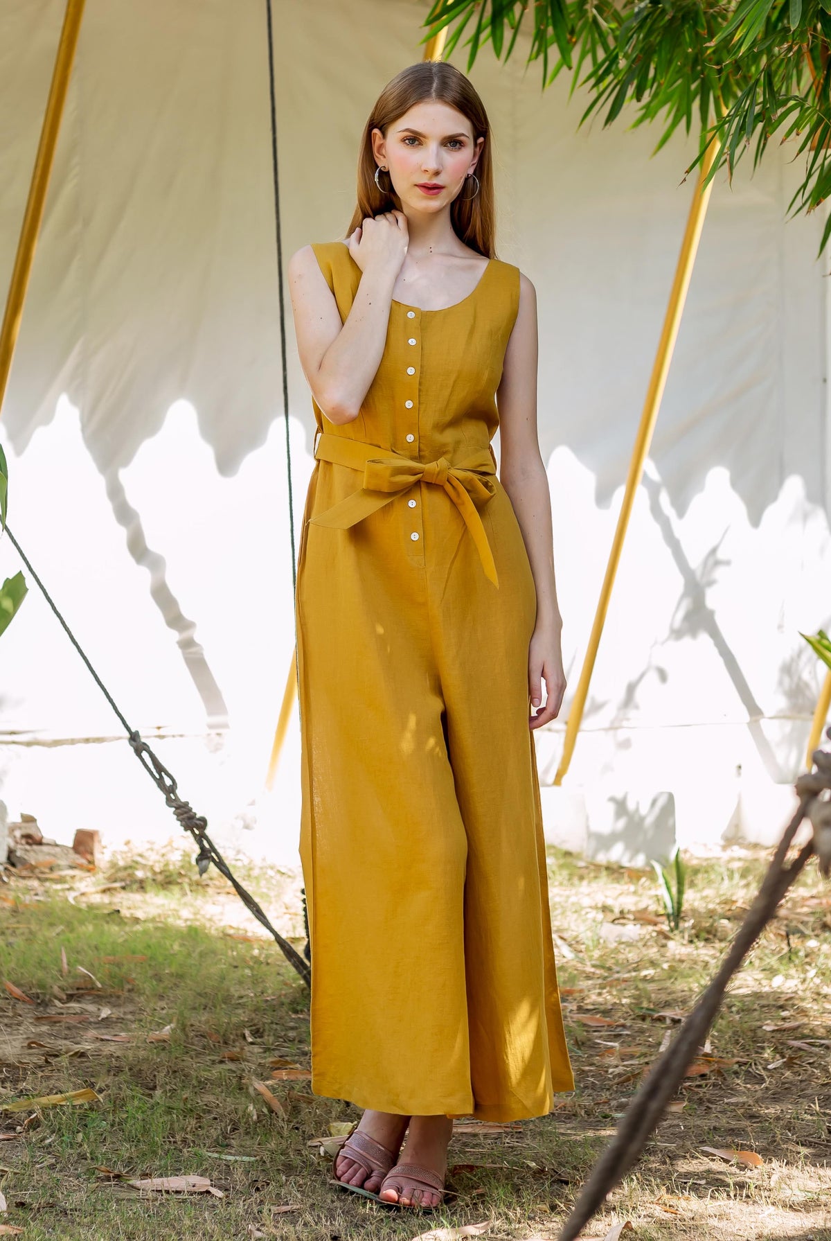 Woman in a sleeveless ochre button-down jumpsuit with a tied waist, standing outdoors.