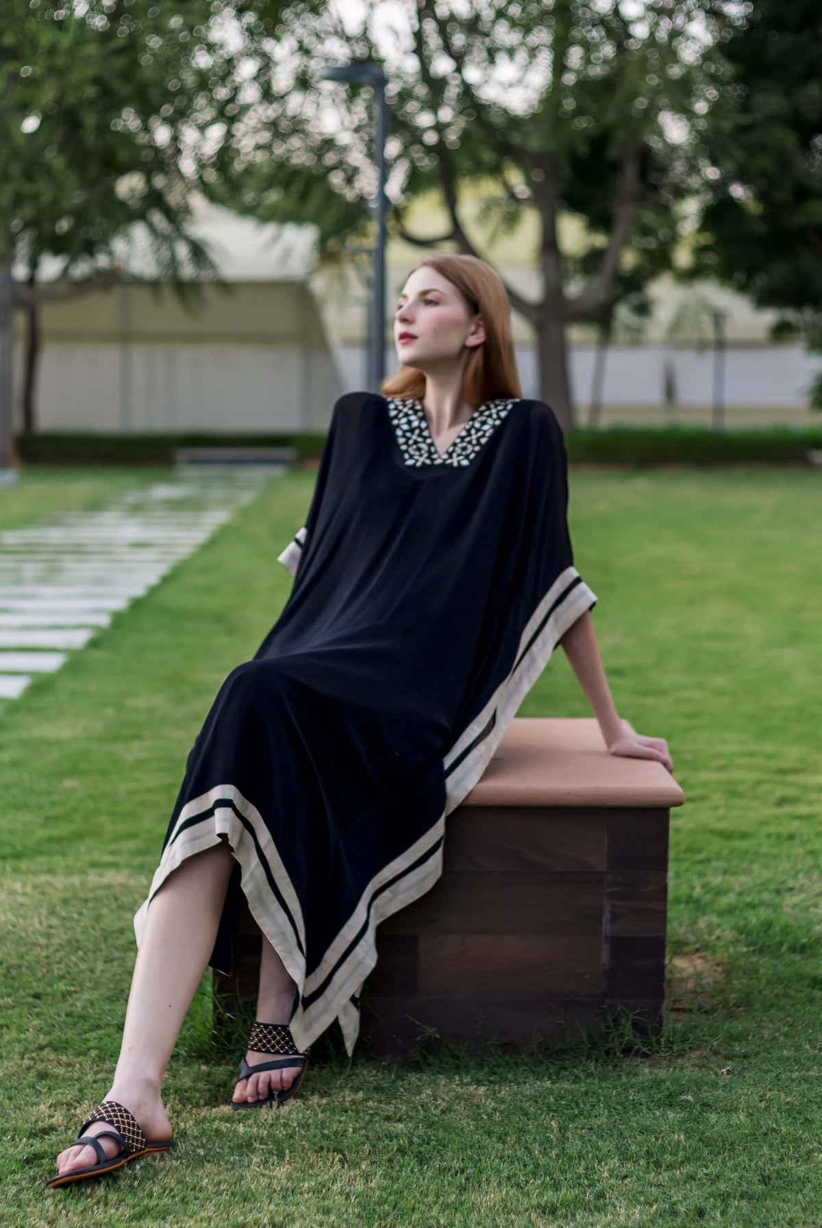 A woman sits on a bench, wearing a black kaftan with white embroidery and a white border.