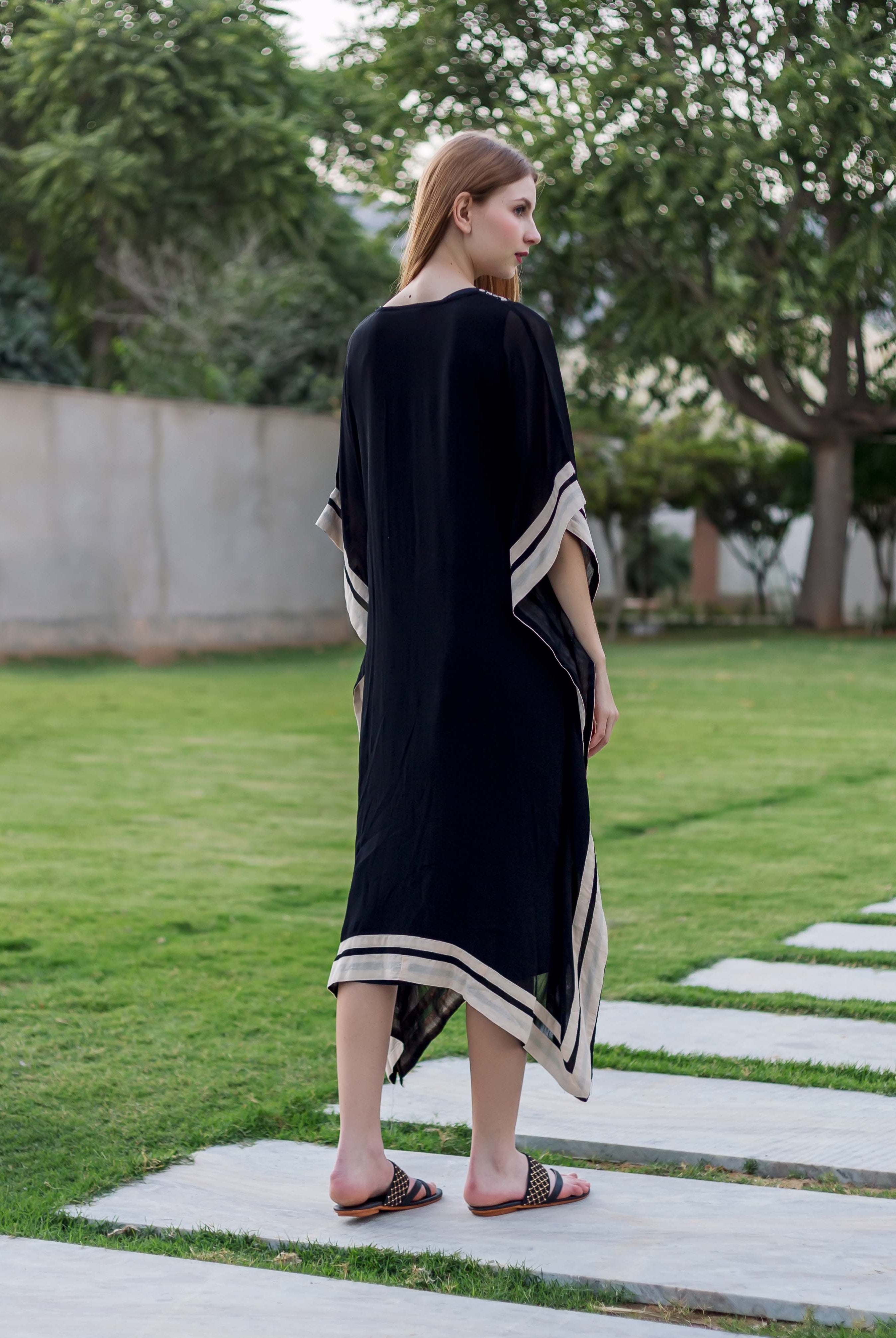 Model from behind wearing a black kaftan with off-white striped borders, standing in a garden.