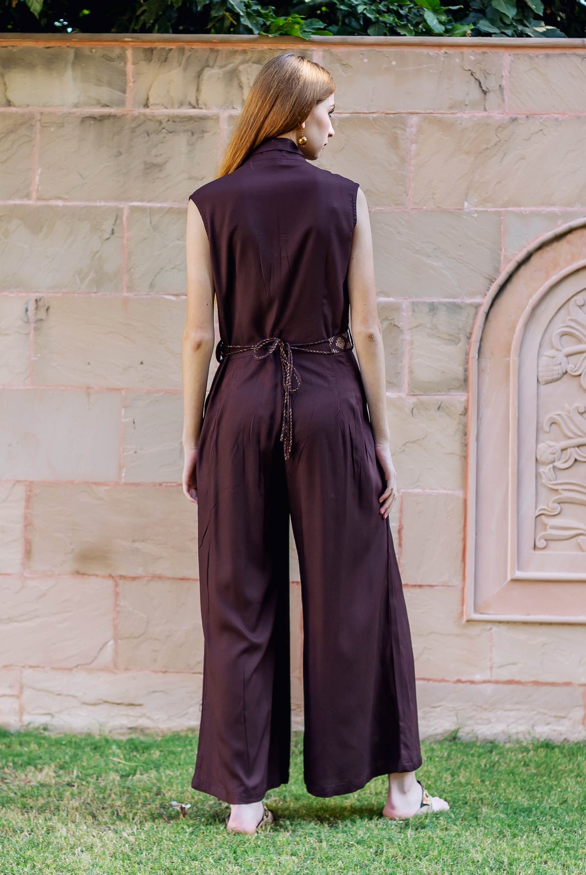 Model from behind in a dark brown sleeveless jumpsuit with braided tie-belt and wide legs.