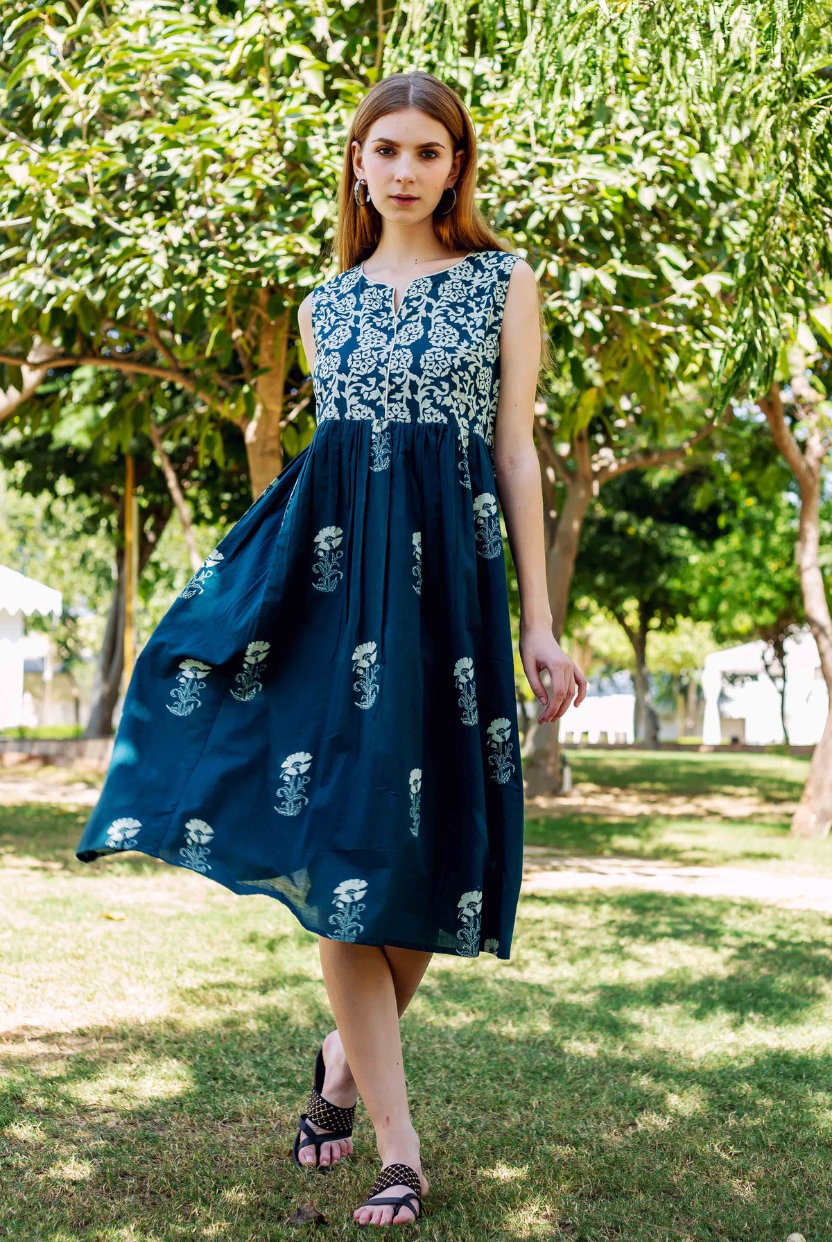 A model wearing a teal green dress with white floral print, standing in a park.