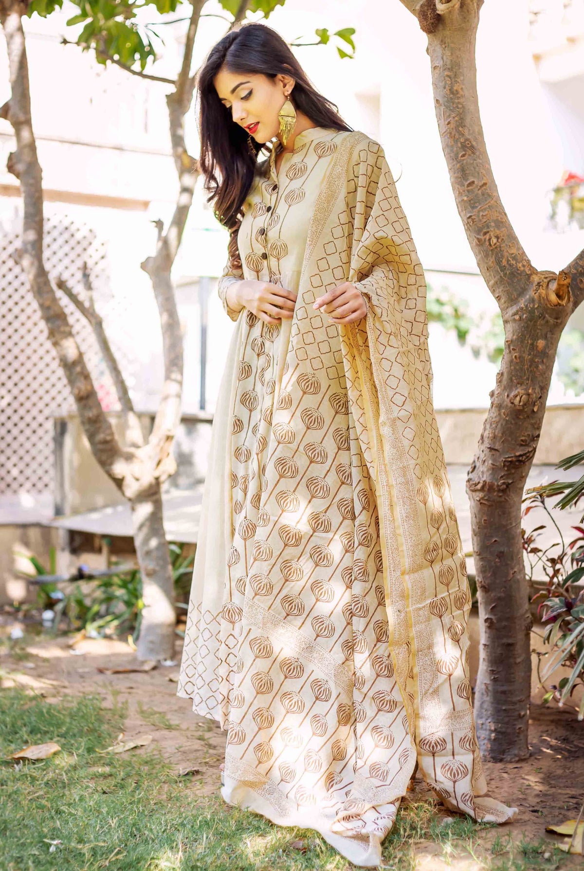 A woman in a beige and brown printed dress stands beside a tree.