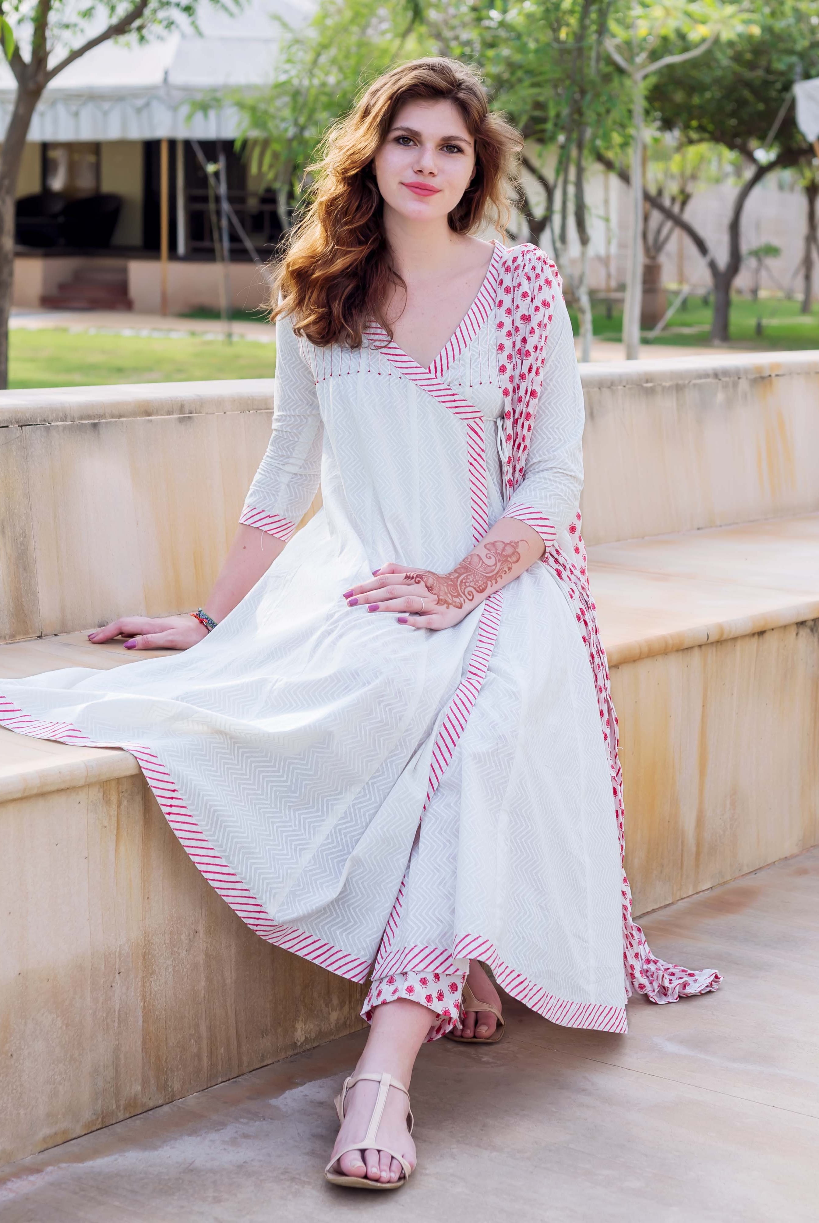 Model wears white Angrakha dress with red hand-block print, sitting on a bench.
