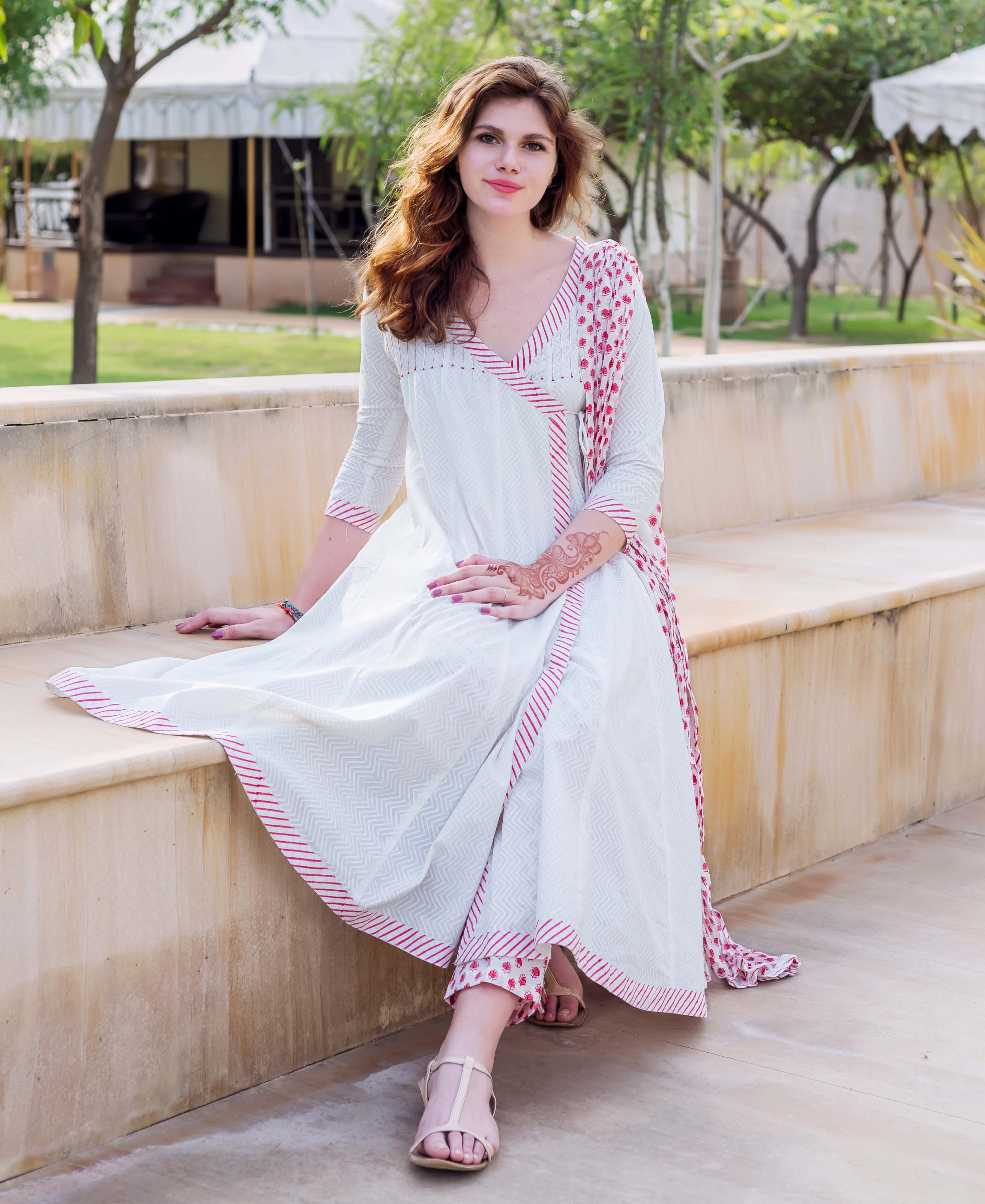 Model wears white Angrakha dress with red hand-block print, sitting on a bench.