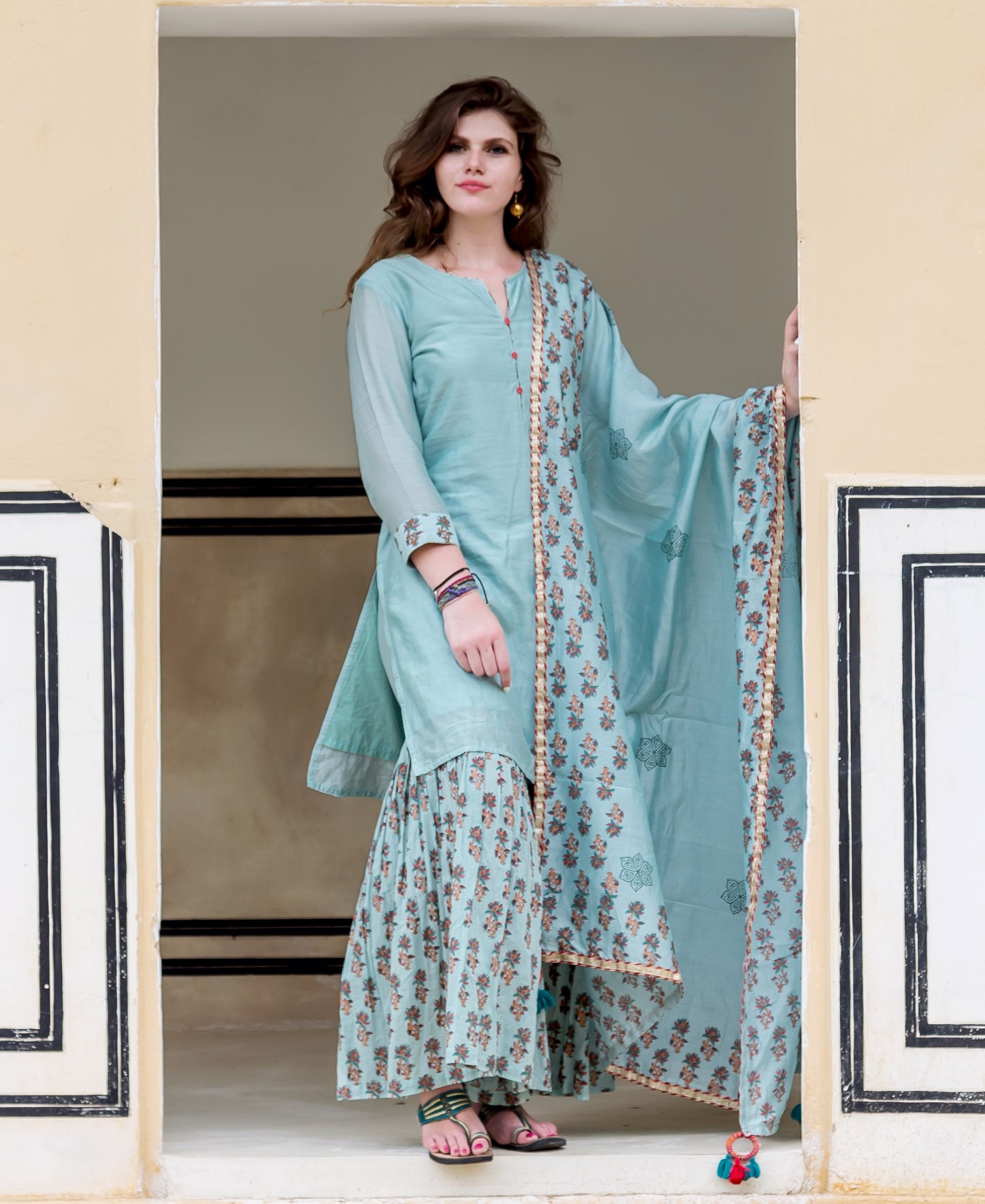 Front full look of model in ice blue solid kurta, floral printed sharara, and dupatta.