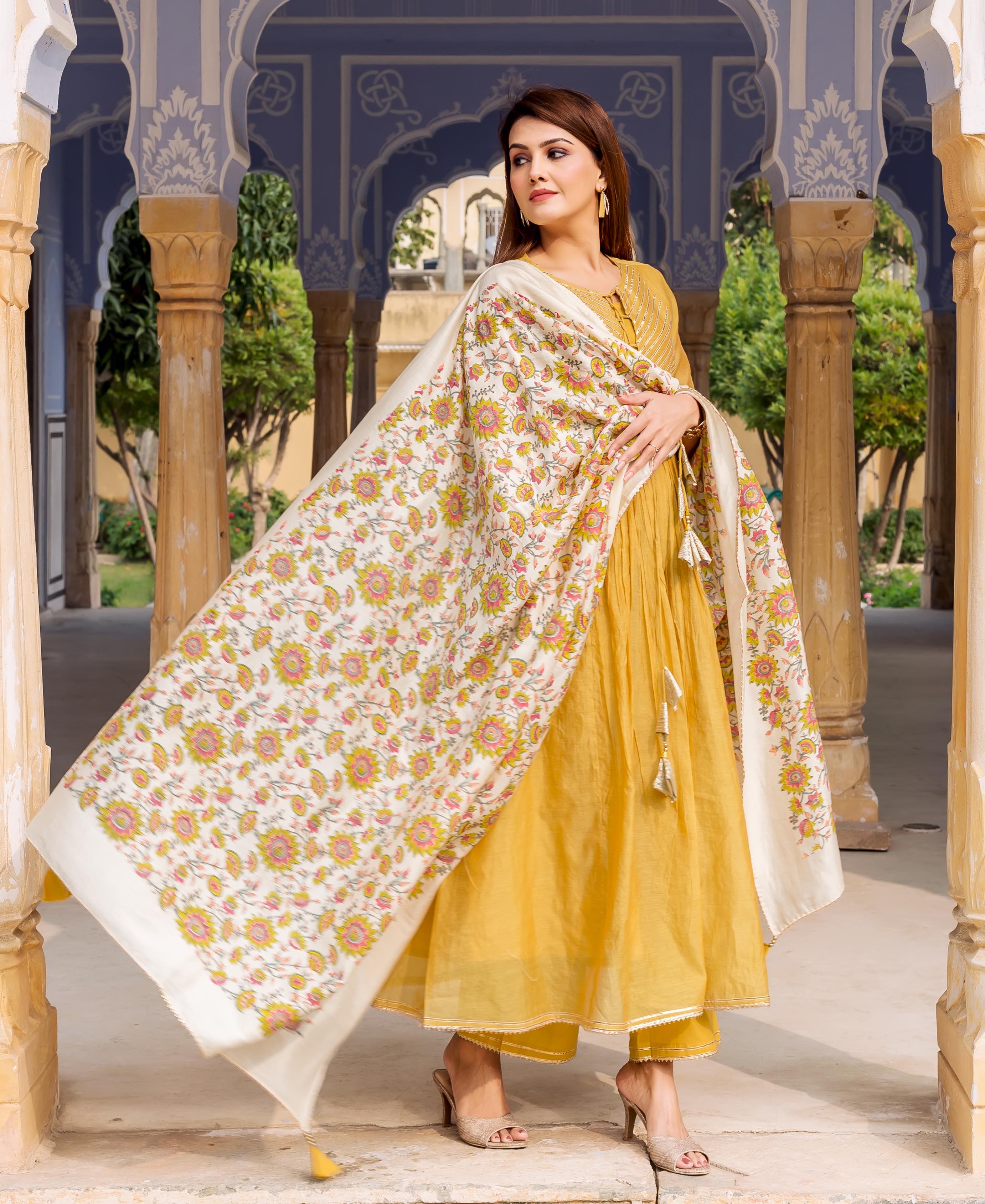 A woman in a yellow kurta with floral print and tassels, posing in an archway.
