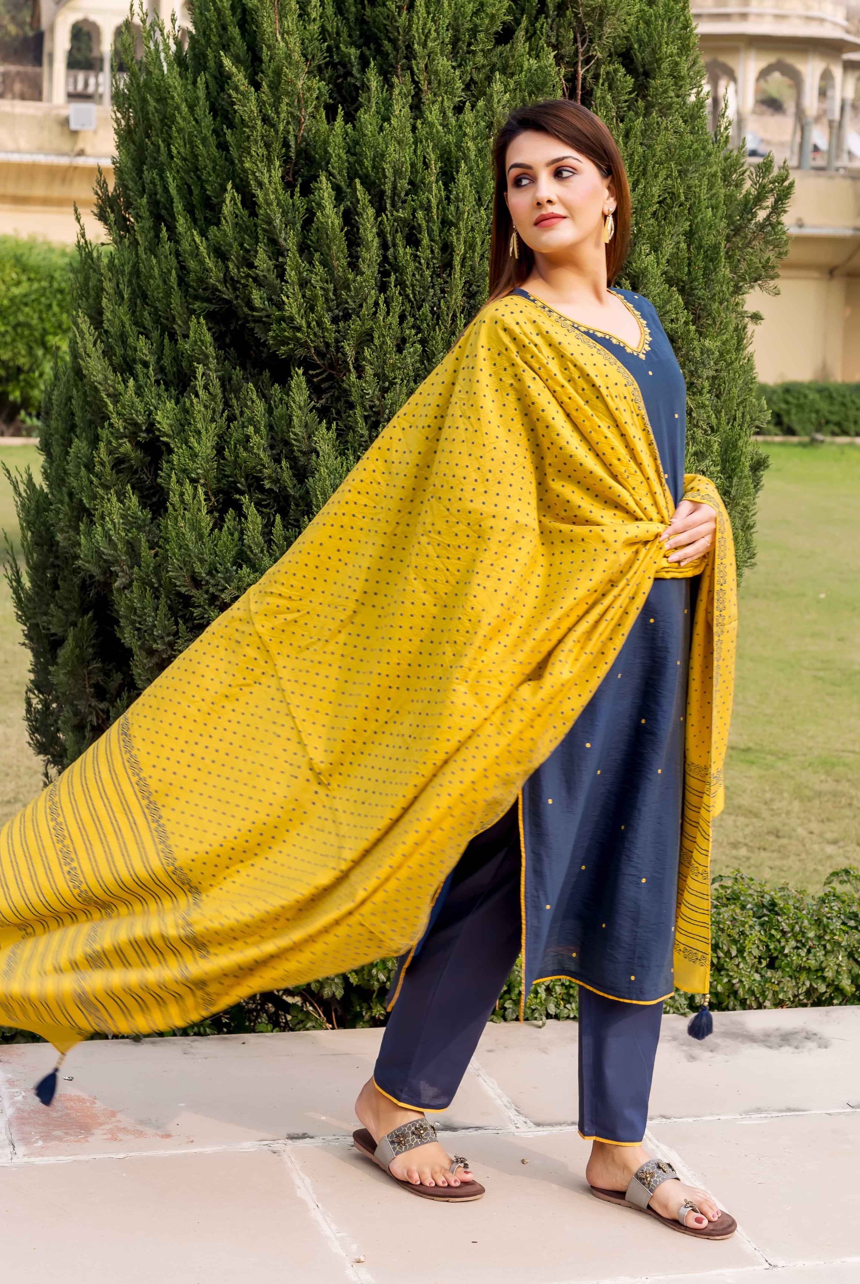 Side pose model wearing navy blue embroidered kurta set with flowing yellow printed dupatta.