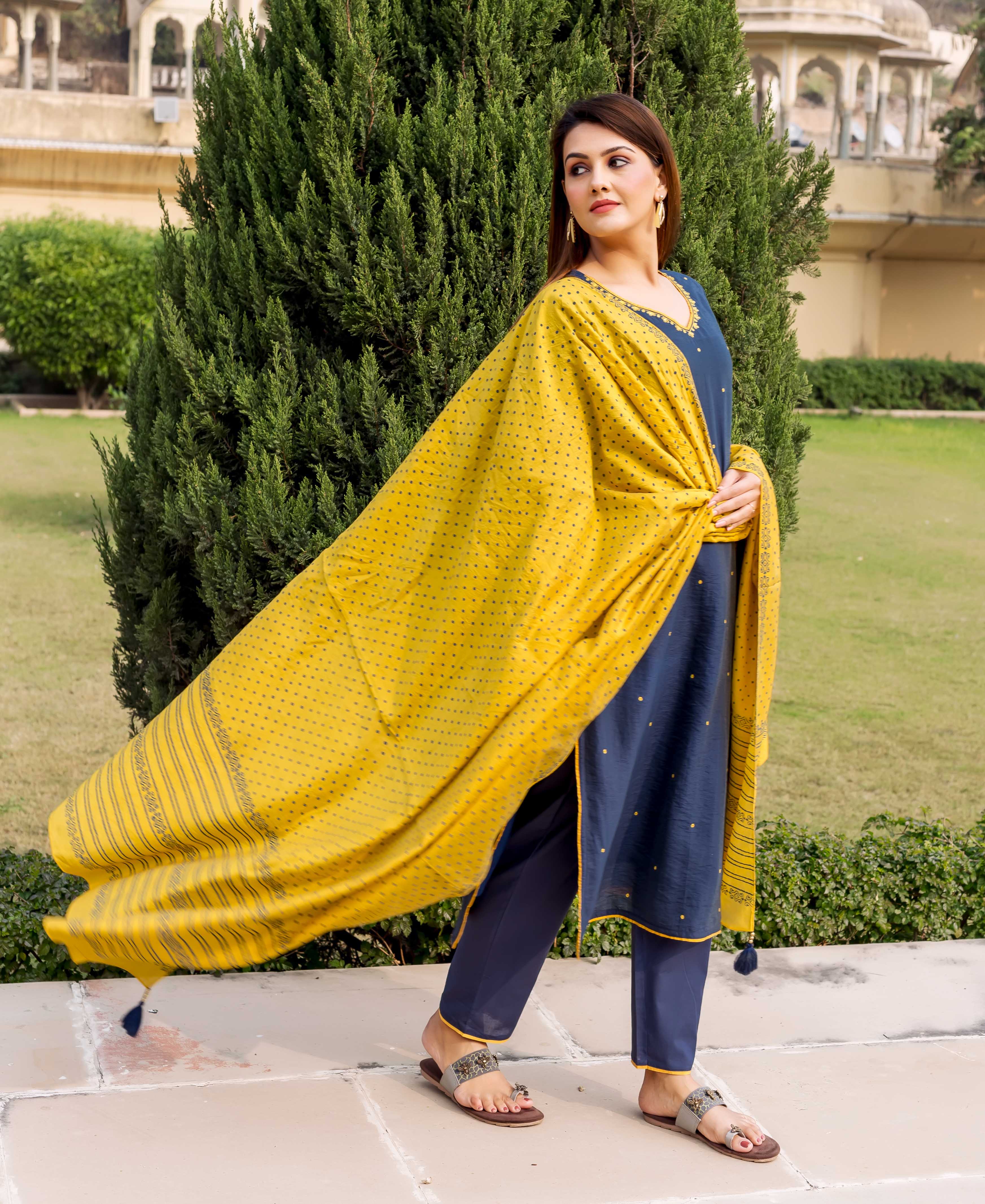 Side pose model wearing navy blue embroidered kurta set with flowing yellow printed dupatta.