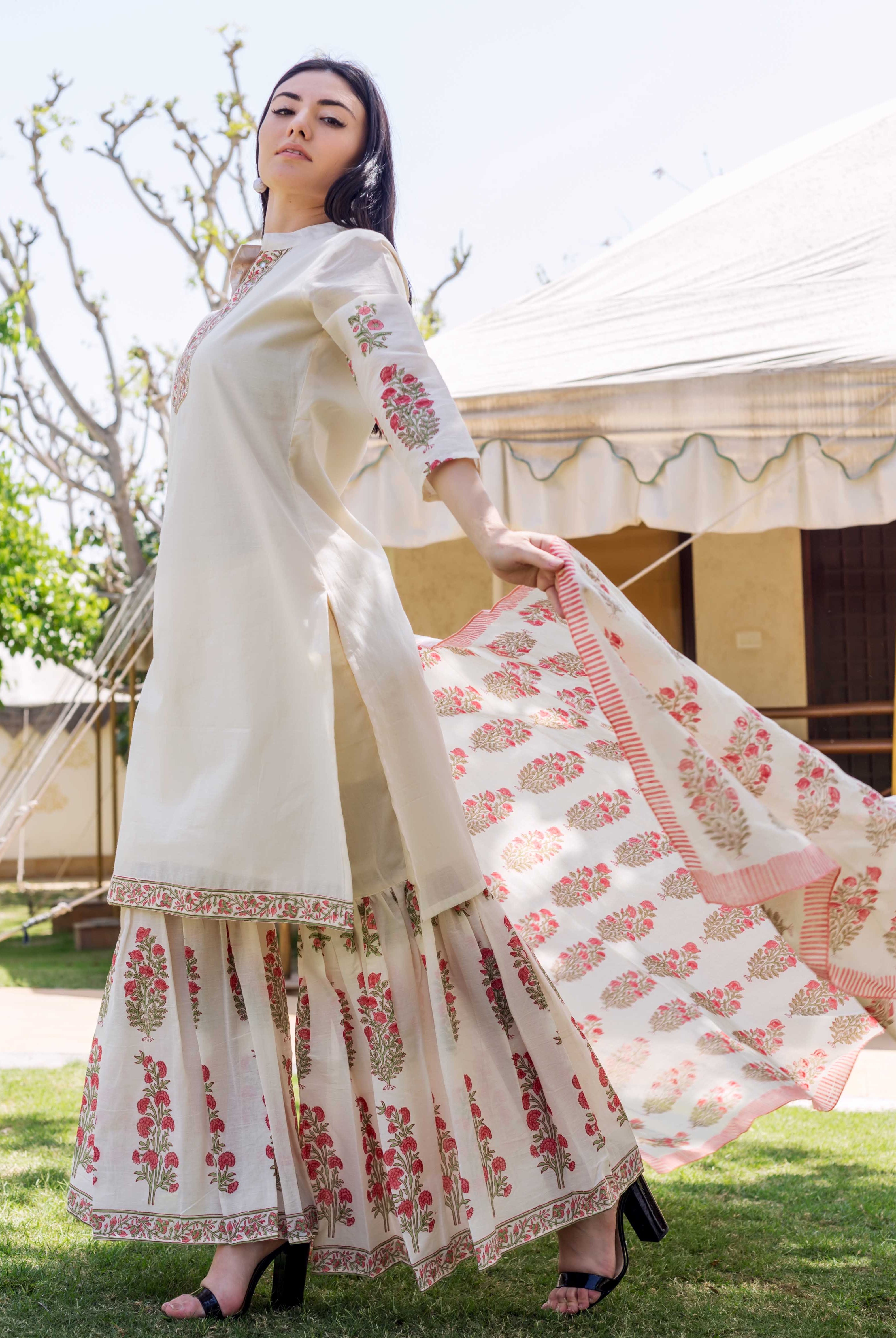 Woman models ivory embroidered kurta, red floral block printed sharara and matching dupatta outdoors.