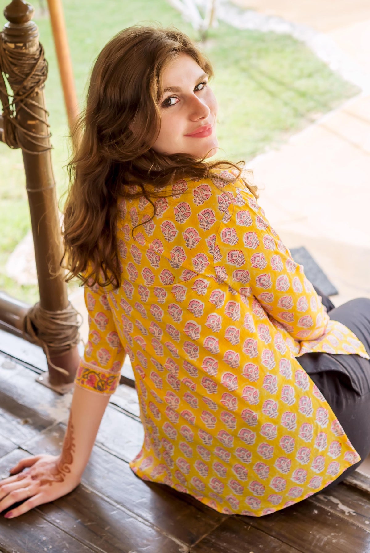 Smiling model wears yellow block-printed top with pink floral motifs, sitting, henna on arm.