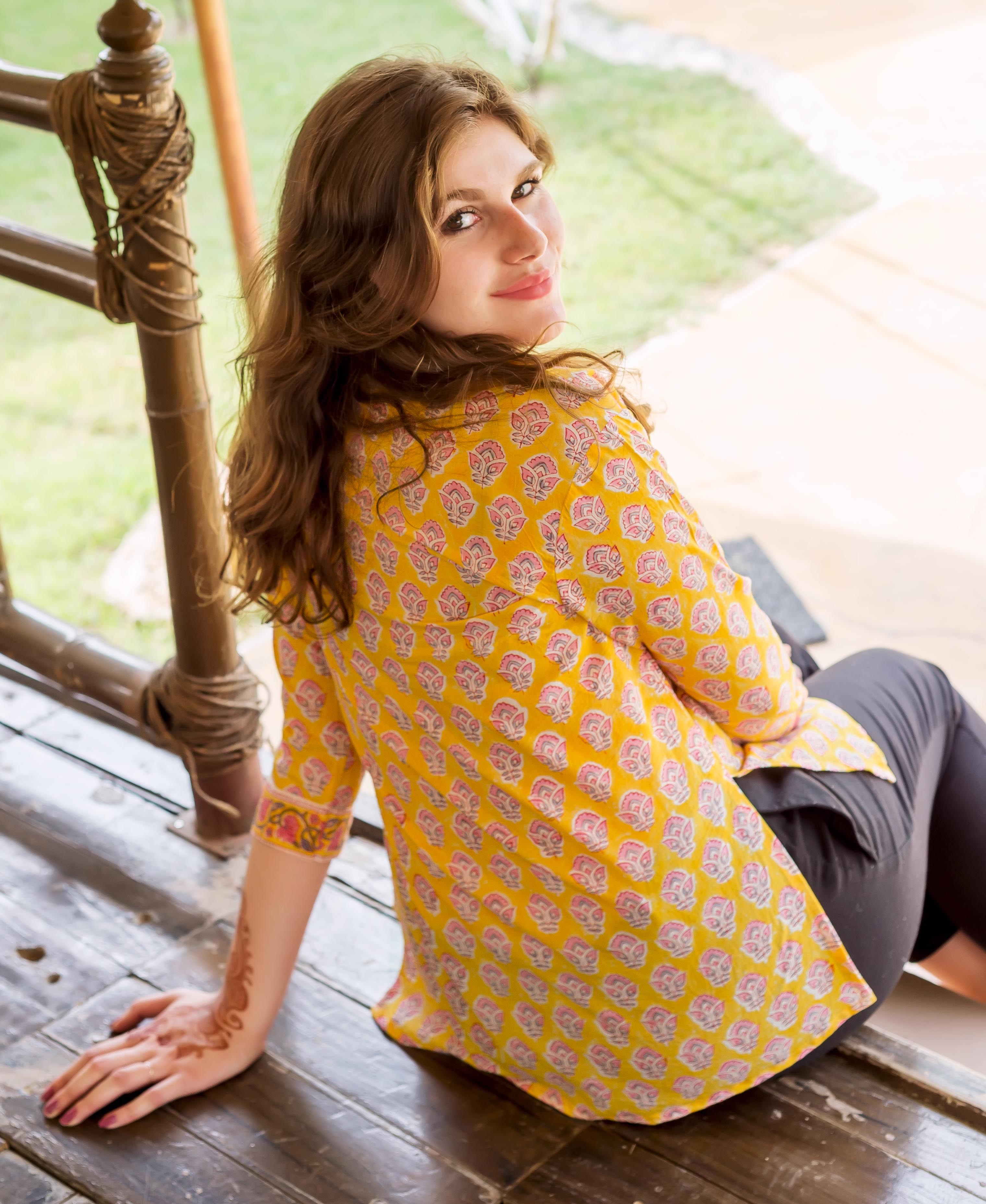 Smiling model wears yellow block-printed top with pink floral motifs, sitting, henna on arm.