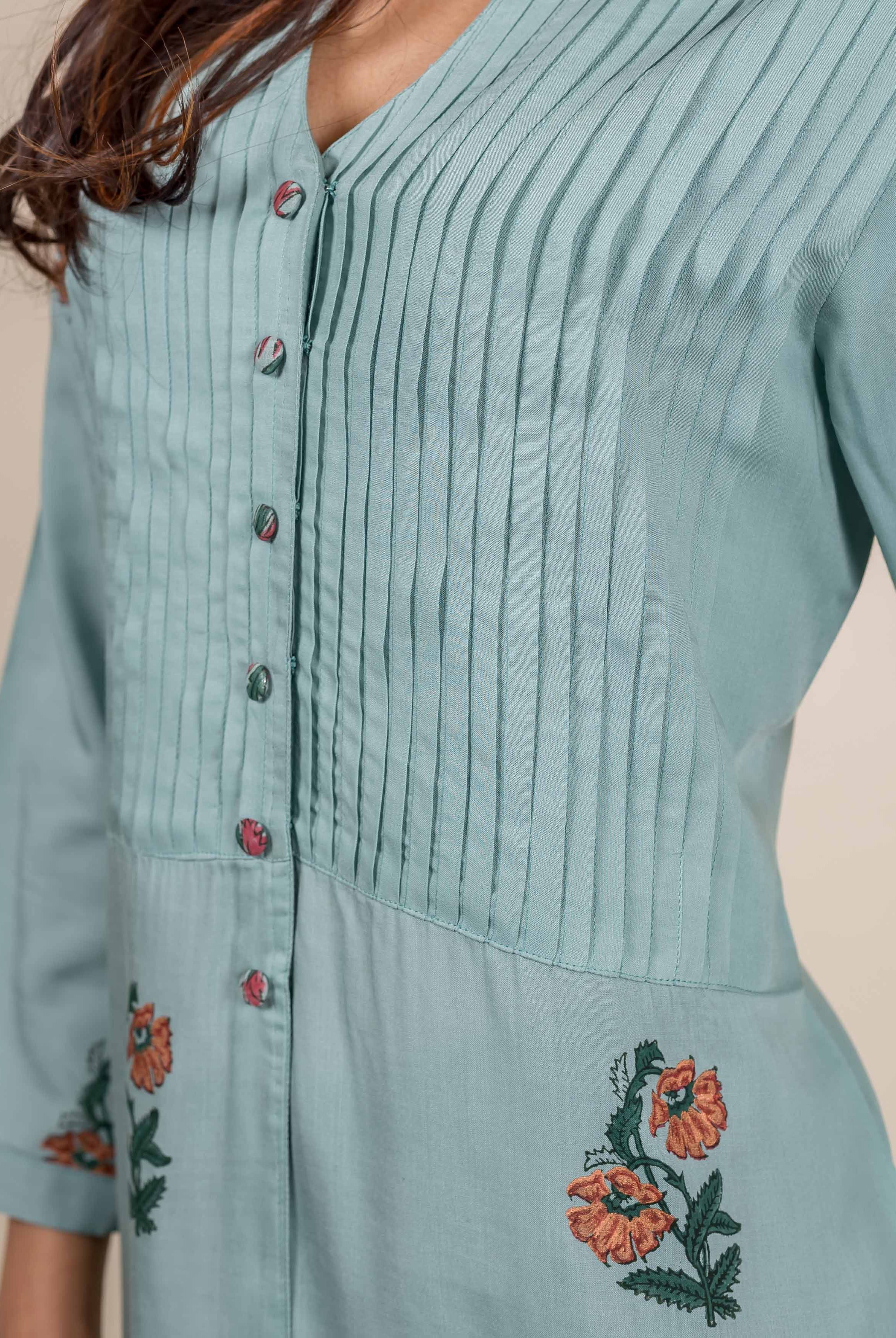 Turquoise blue top with pin tucks and orange floral embroidery on a pale background.