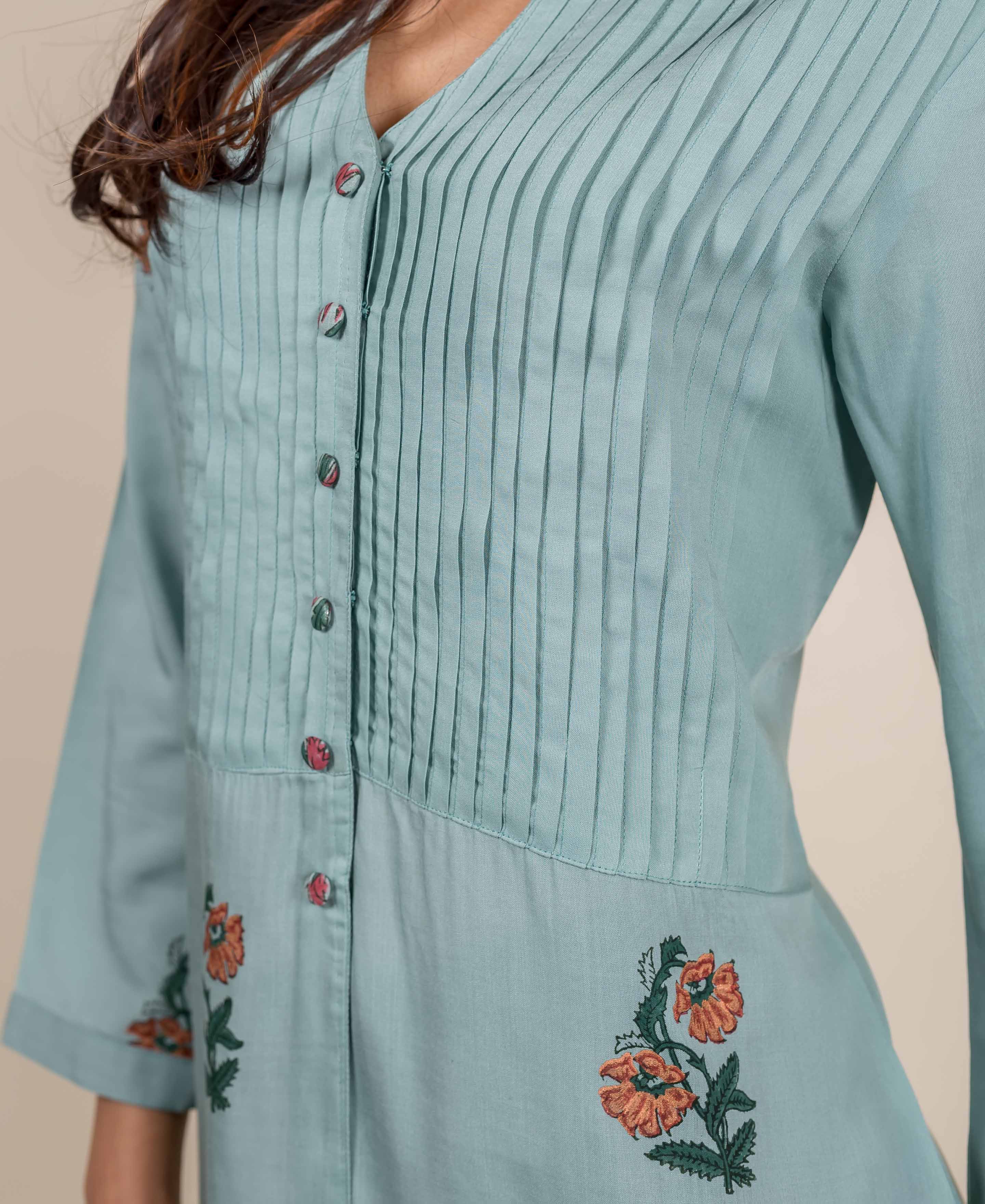 Turquoise blue top with pin tucks and orange floral embroidery on a pale background.