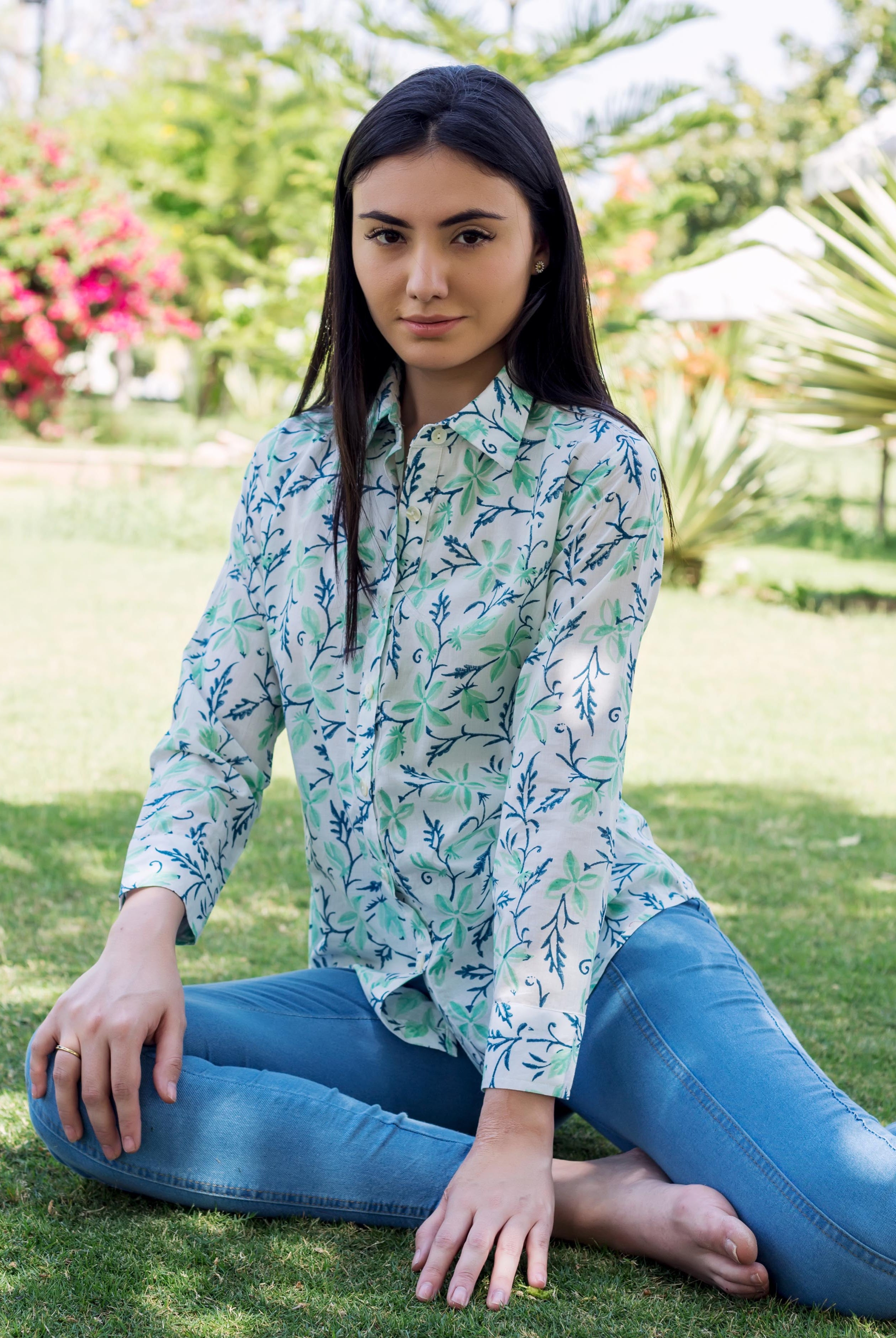 Model wearing sea green and blue block printed shirt and blue jeans, sitting outdoors.