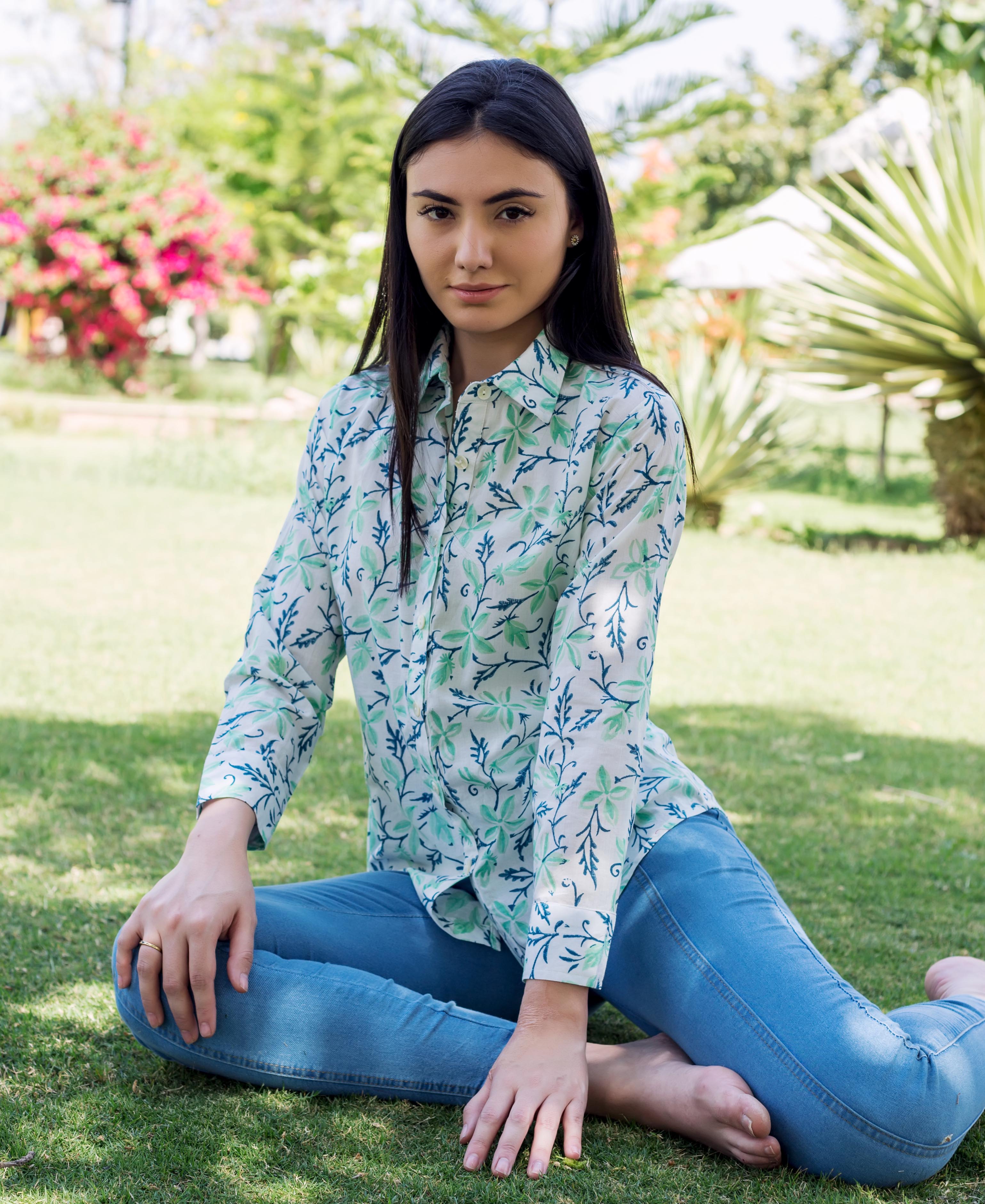 Model wearing sea green and blue block printed shirt and blue jeans, sitting outdoors.