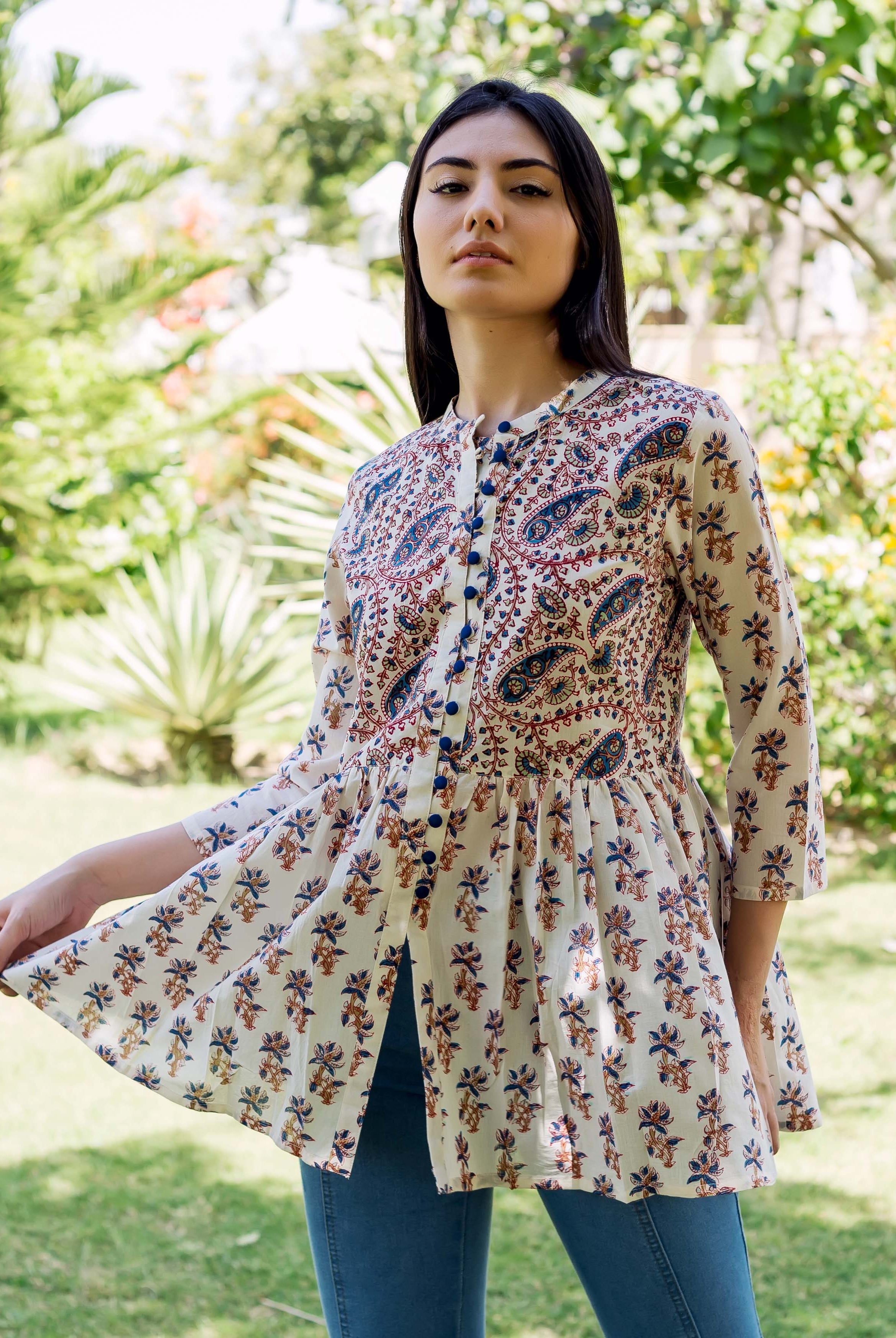 Model wearing a cream peplum top with blue, red, and brown ethnic patterns in a garden.